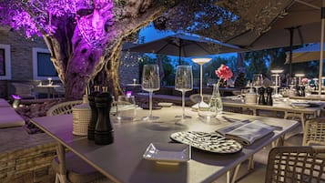 Un espacio de comedor al aire libre con mesas, sillas y un árbol con flores moradas.