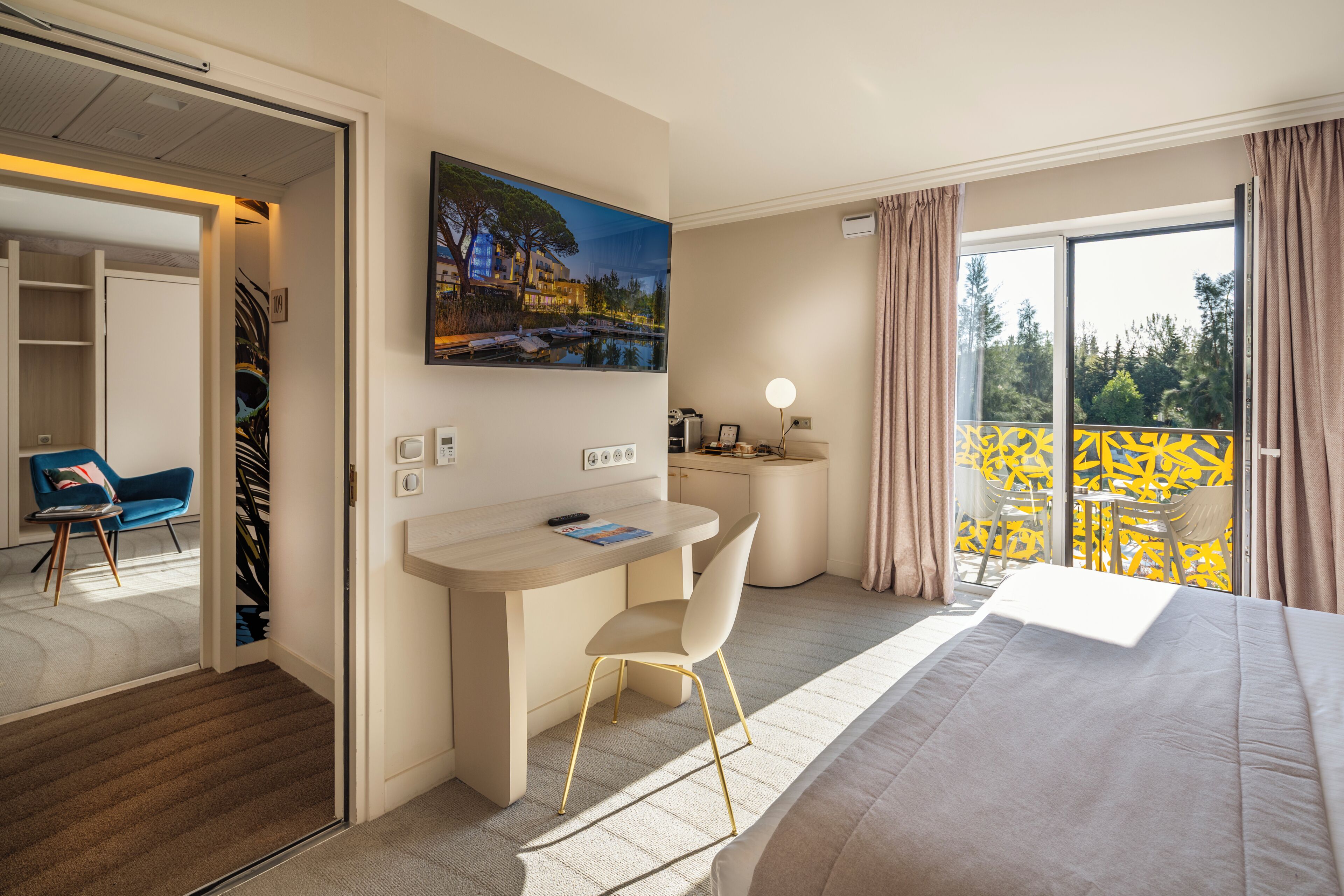 Ein Hotelzimmer mit Bett, Schreibtisch, Stuhl, einem Gemälde und einem Balkon mit gelber Geländer.