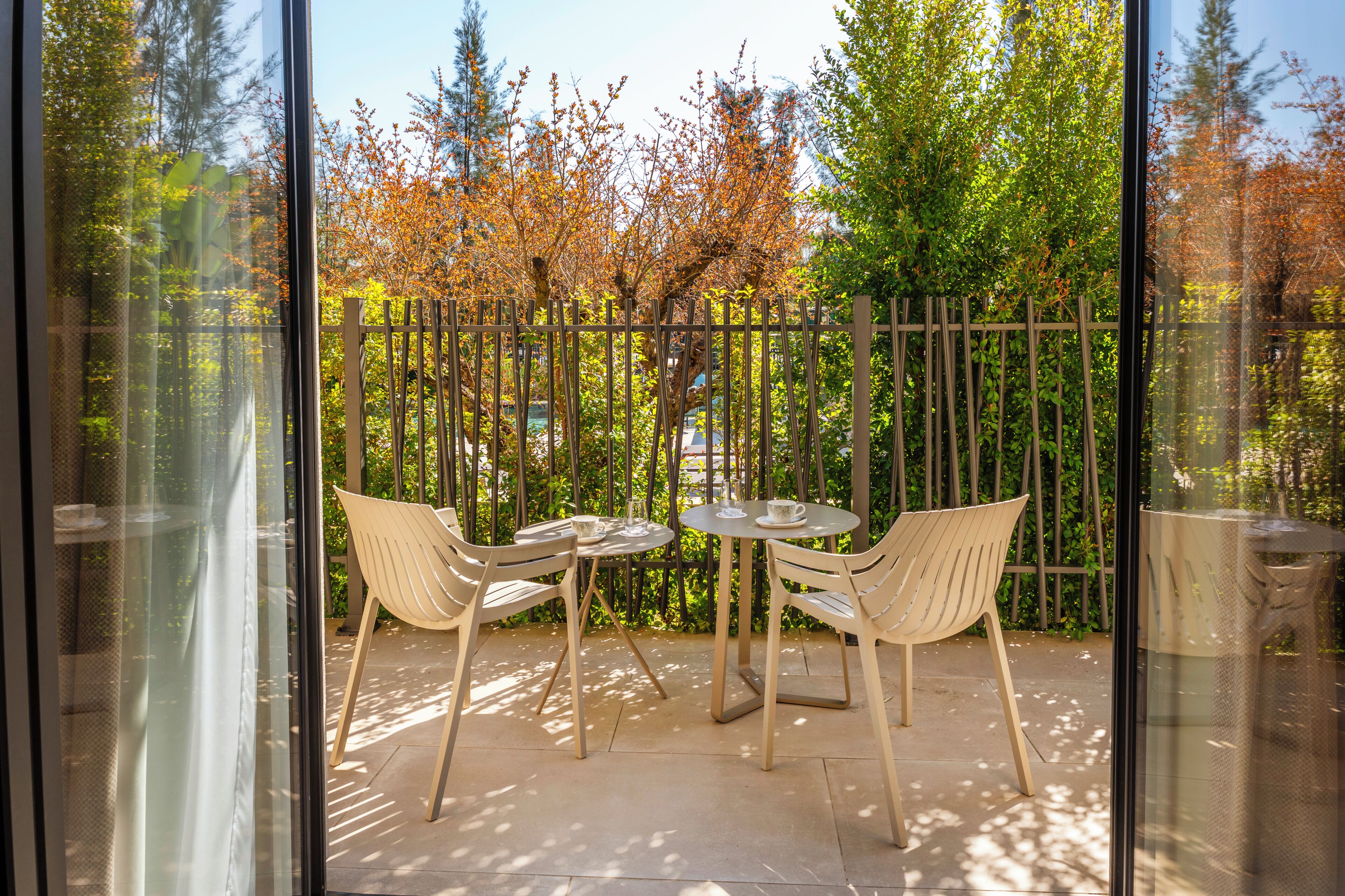 A patio with two chairs and a table, a glass wall, and a view of trees and a fence.