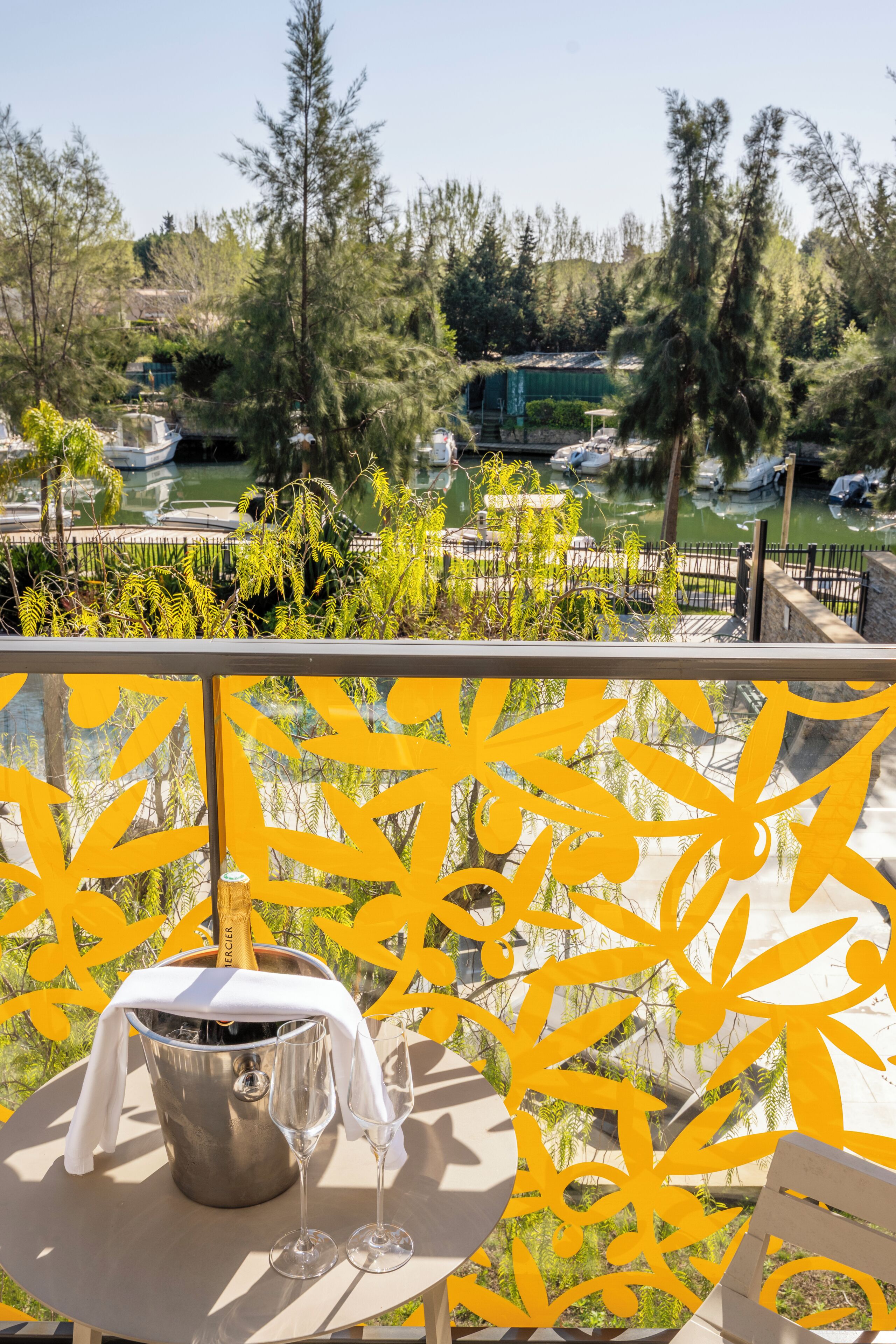 A balcony with a view of a marina, a metal bucket with a bottle, and two wine glasses.