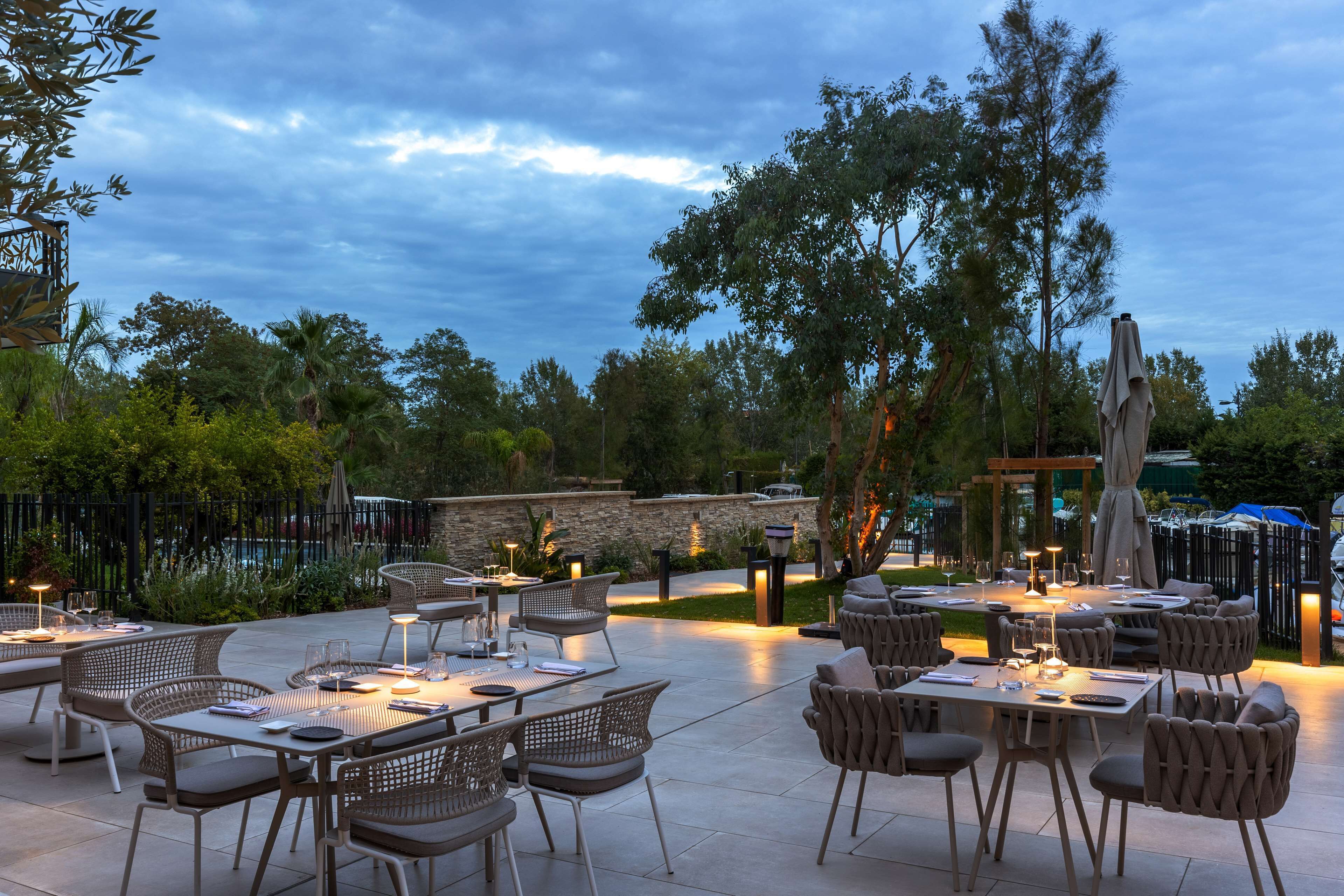 An outdoor dining area with tables and chairs, surrounded by trees and a stone wall.