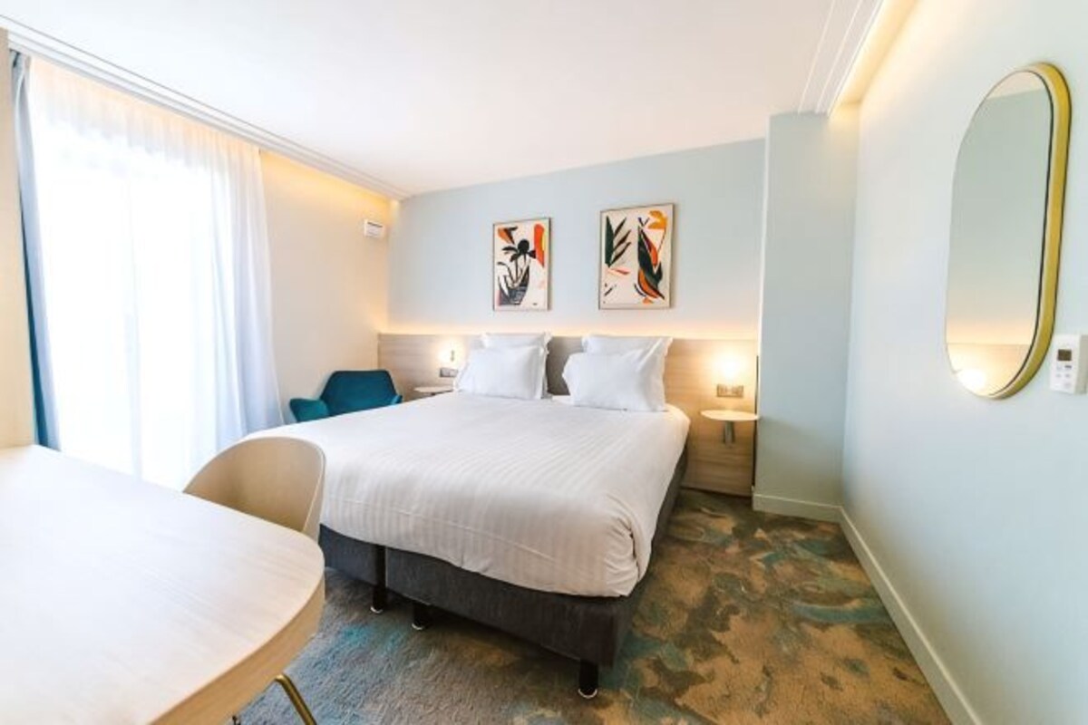 A hotel room with a bed, a desk, a chair, a large mirror, and artwork on the wall.