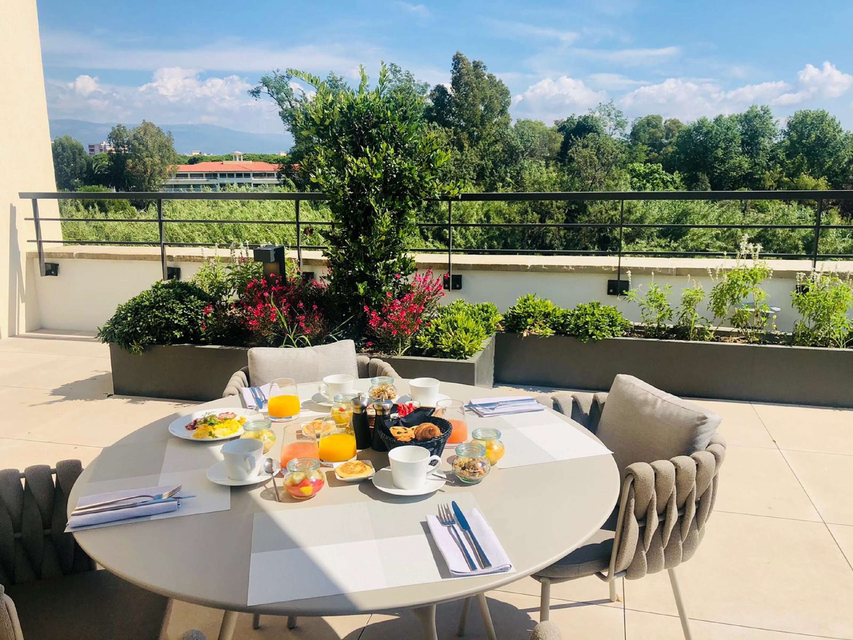 A round table set for breakfast on a terrace with a view of greenery and buildings in the background.