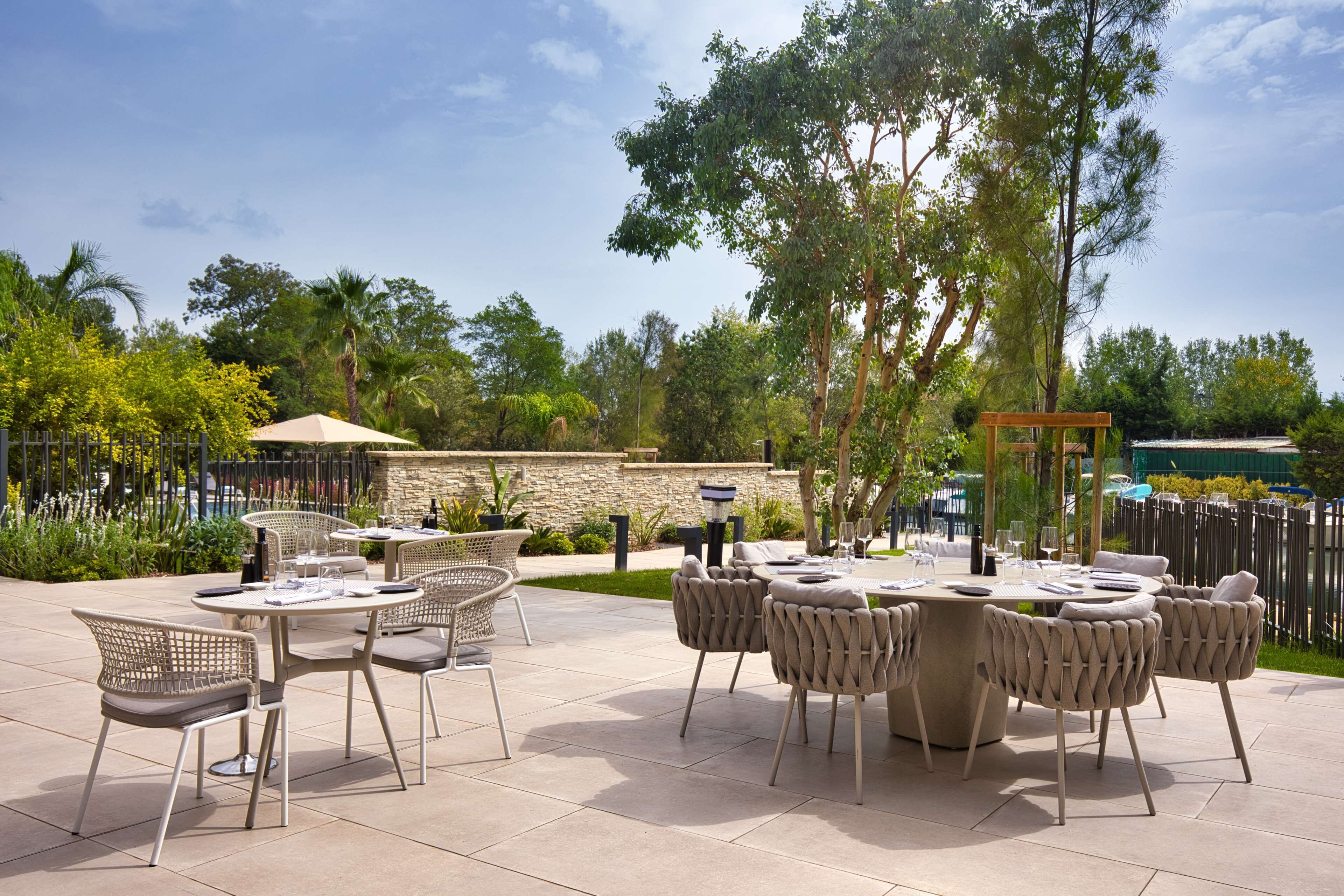 An outdoor patio with round tables and wicker chairs, surrounded by greenery and a stone wall.