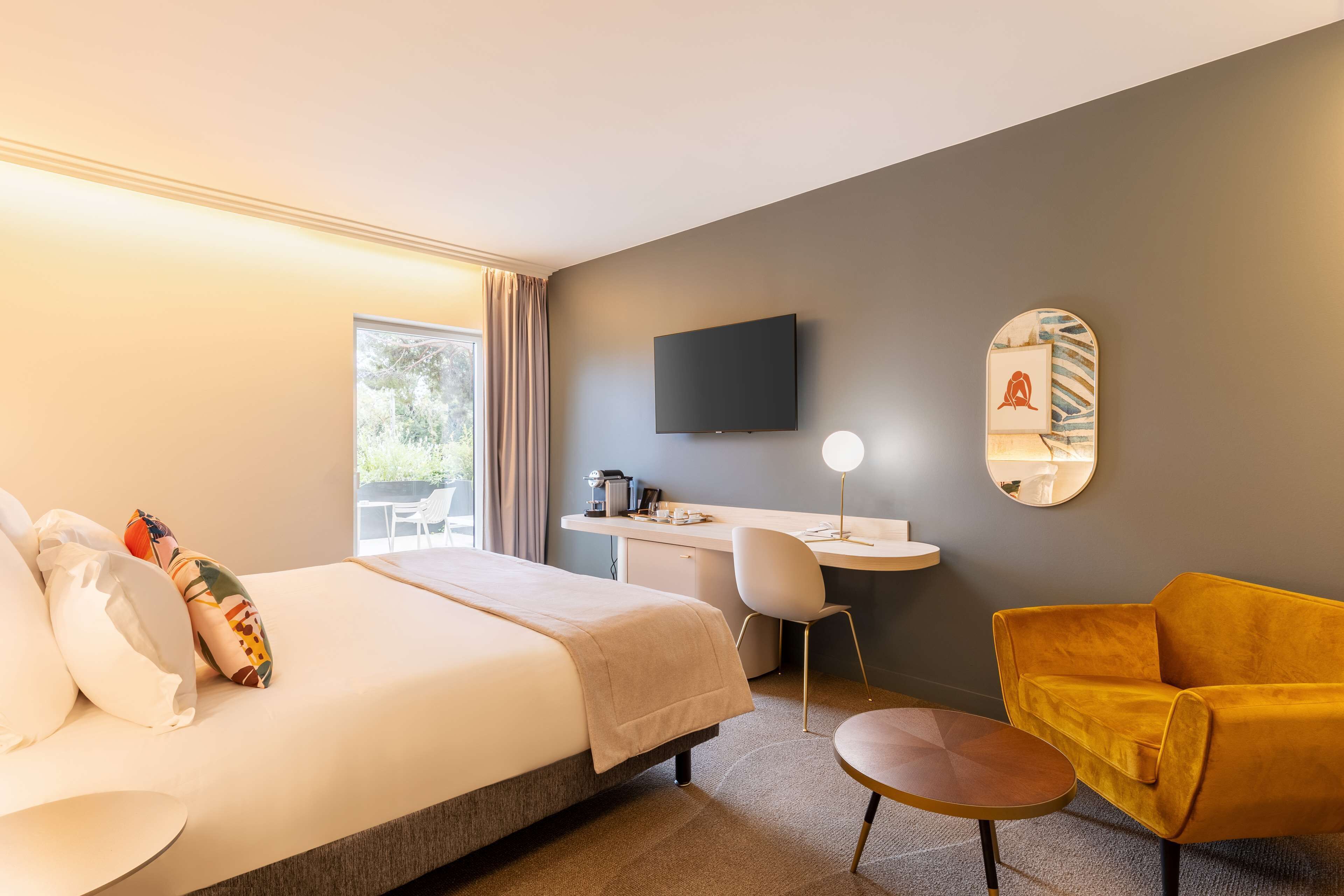 A hotel room with a bed, desk, chair, and a yellow armchair.