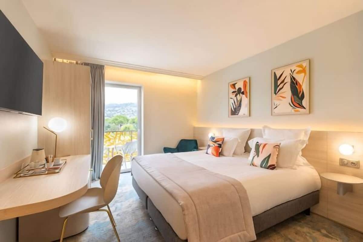 A hotel room with a bed, bedside table, chair, desk, and artwork on the walls.