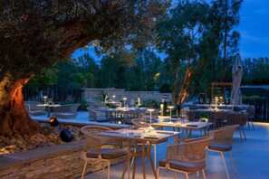 An outdoor dining area with multiple tables and chairs, surrounded by trees and a stone wall.