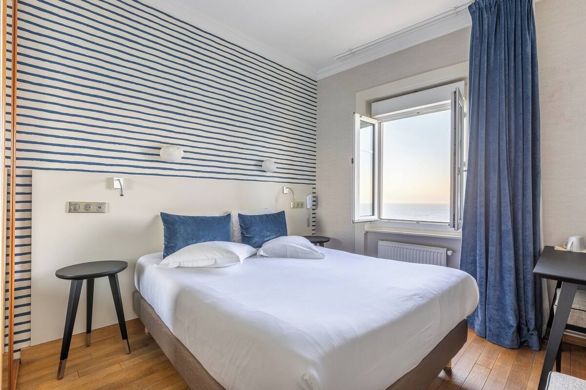 Ein Hotelzimmer mit einem großen Bett, Meerblick durch ein offenes Fenster, einem kleinen Tisch und einem Stuhl.