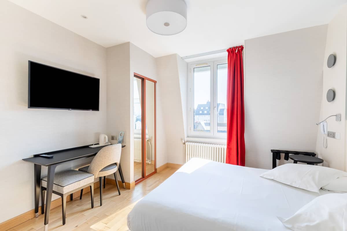 Ein Hotelzimmer mit einem Bett, einem Schreibtisch mit Stuhl, einem Fernseher und einem Fenster mit roten Vorhängen.