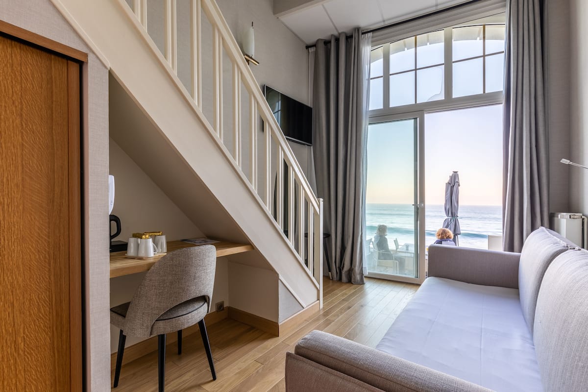 Ein modernes Hotelzimmer mit Schreibtisch aus Holz, einer gemütlichen Couch und einer Treppe, die auf einen Balkon mit Meerblick führt.