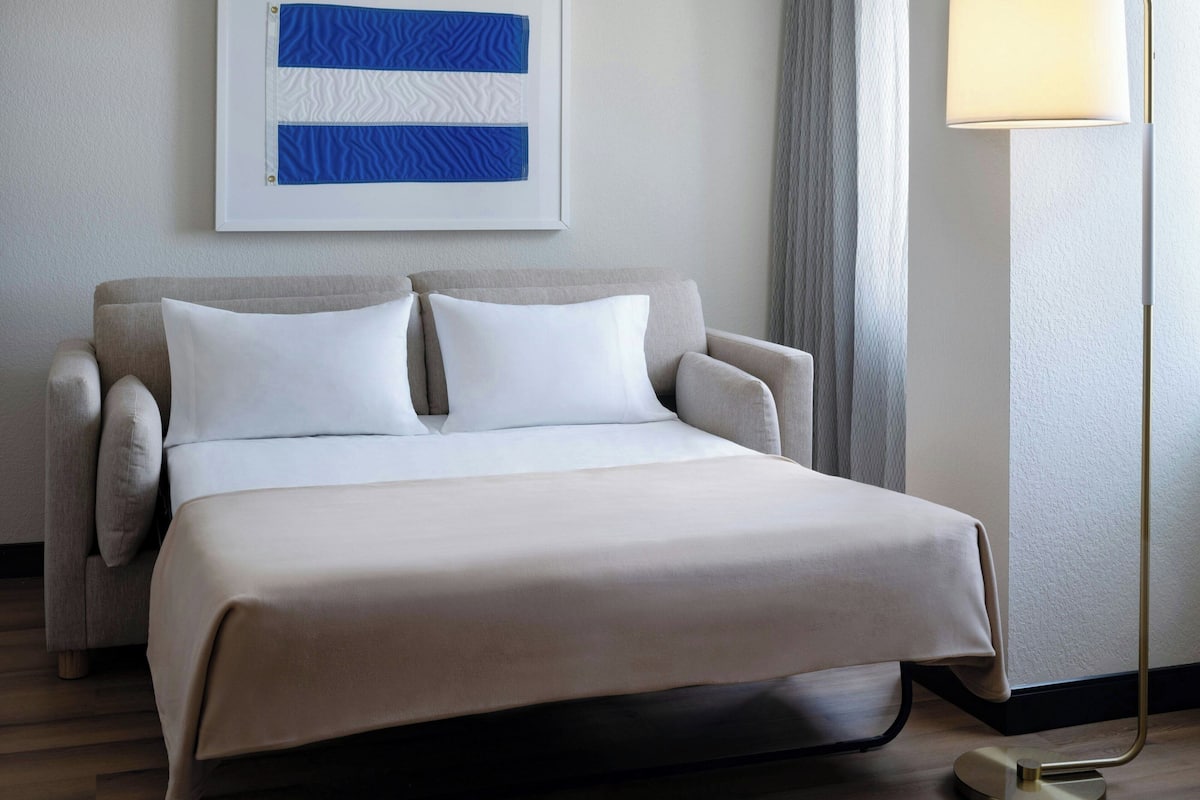 Una cama bien hecha con colcha y almohadas blancas, un sofá gris con cojines a juego, una obra de arte abstracta enmarcada en la pared y una lámpara de pie junto a la cama.