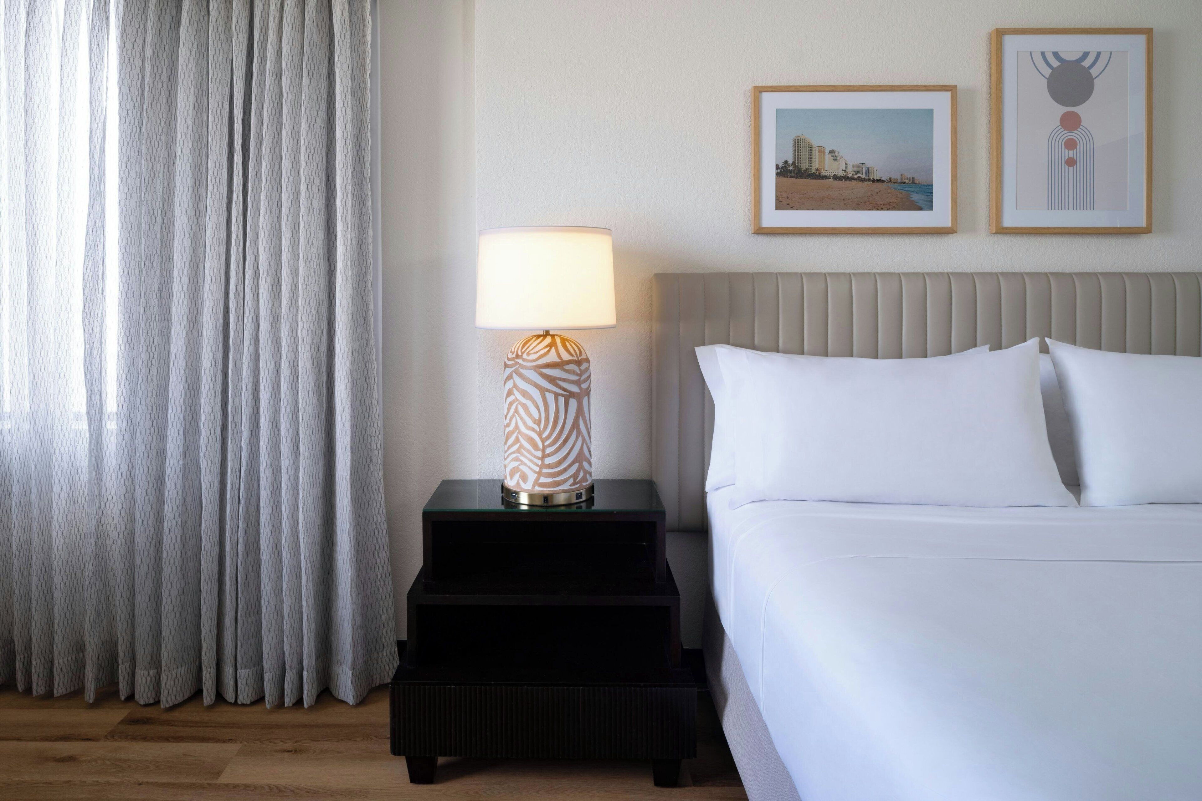 Une chambre d’hôtel avec un lit, une table de chevet avec une lampe, un tableau encadré et une fenêtre avec des rideaux.