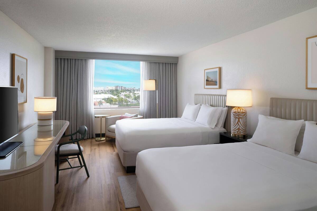 Habitación de hotel con dos camas, un escritorio con televisor, una silla y vistas a la ciudad.