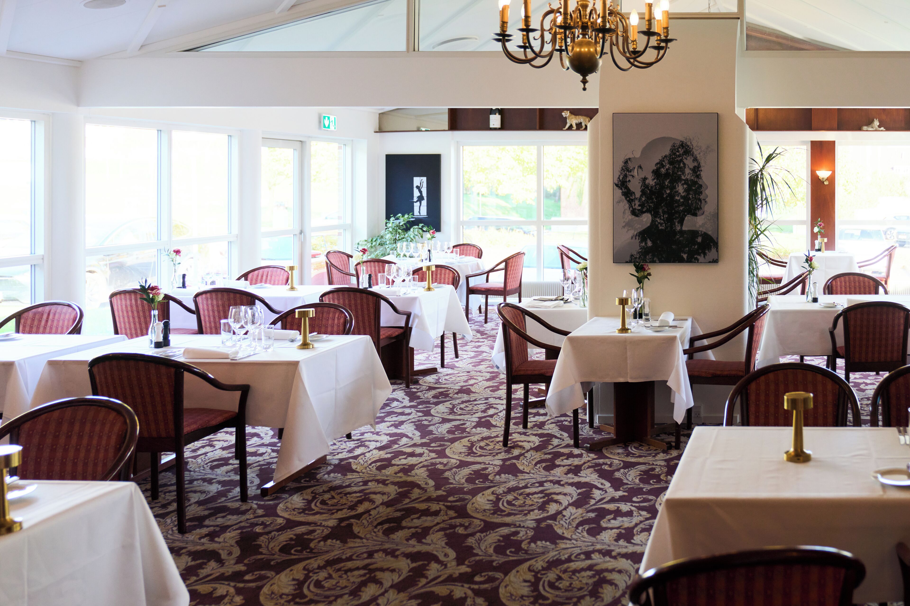 A restaurant with multiple tables and chairs, large windows, and a chandelier.