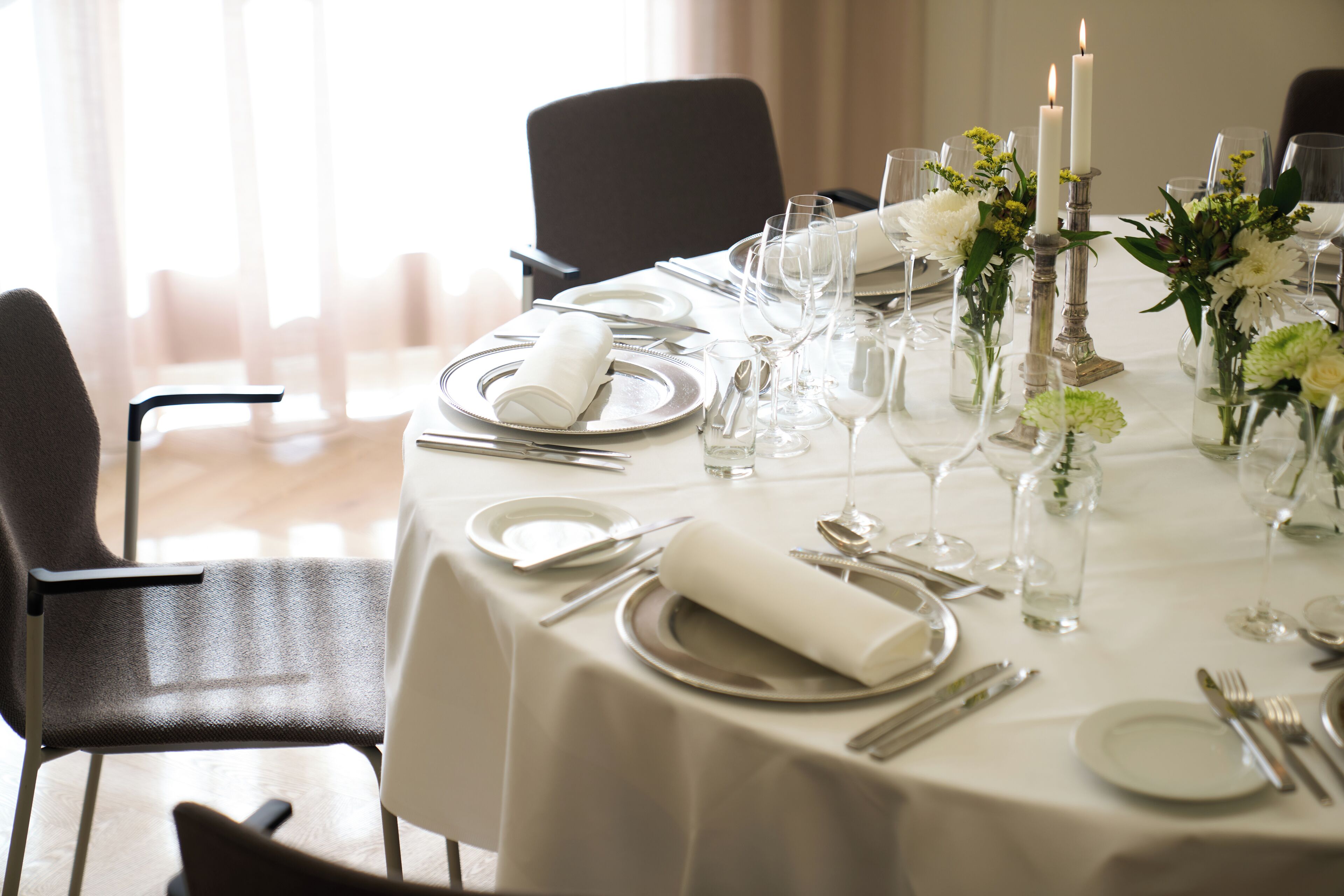 Ein runder Esstisch ist für ein festliches Essen gedeckt, mit weißer Tischdecke, Besteck, Tellern und Gläsern.