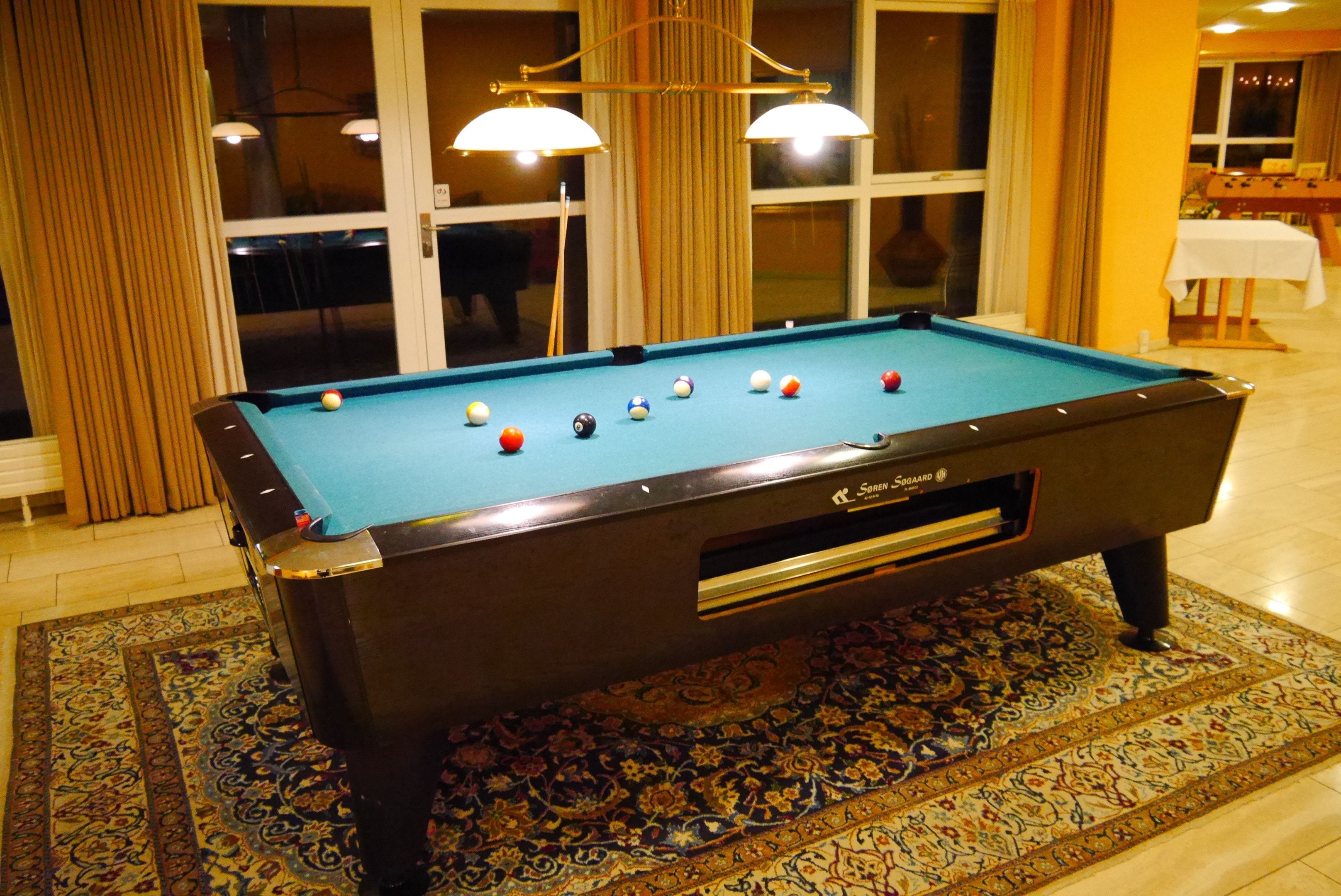 A pool table with balls, a patterned rug, and a window with curtains.