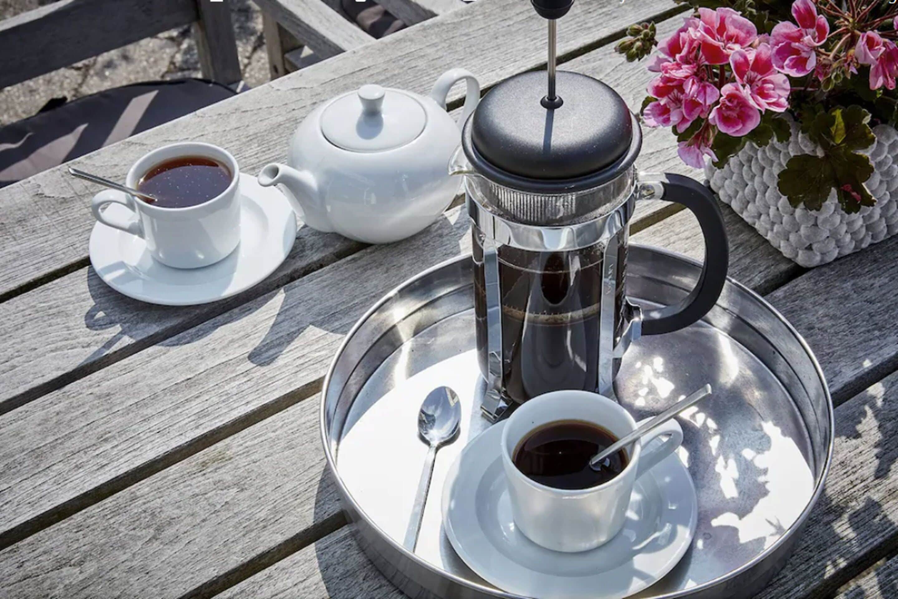 Una bandeja metálica con una prensa francesa, una tetera y dos tazas de café sobre una mesa de madera.