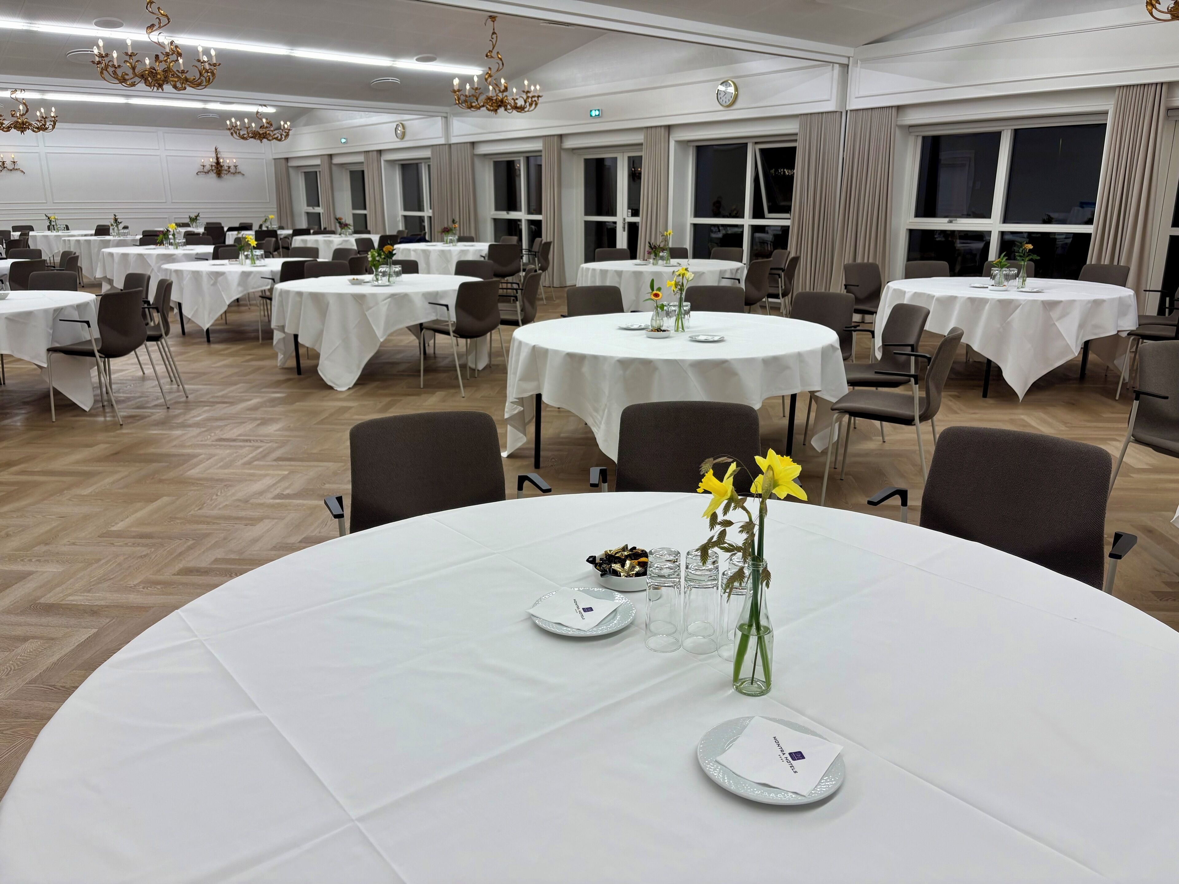 Un salón de banquetes con varias mesas redondas, cada una con mantel blanco, sillas y pequeños centros de mesa.