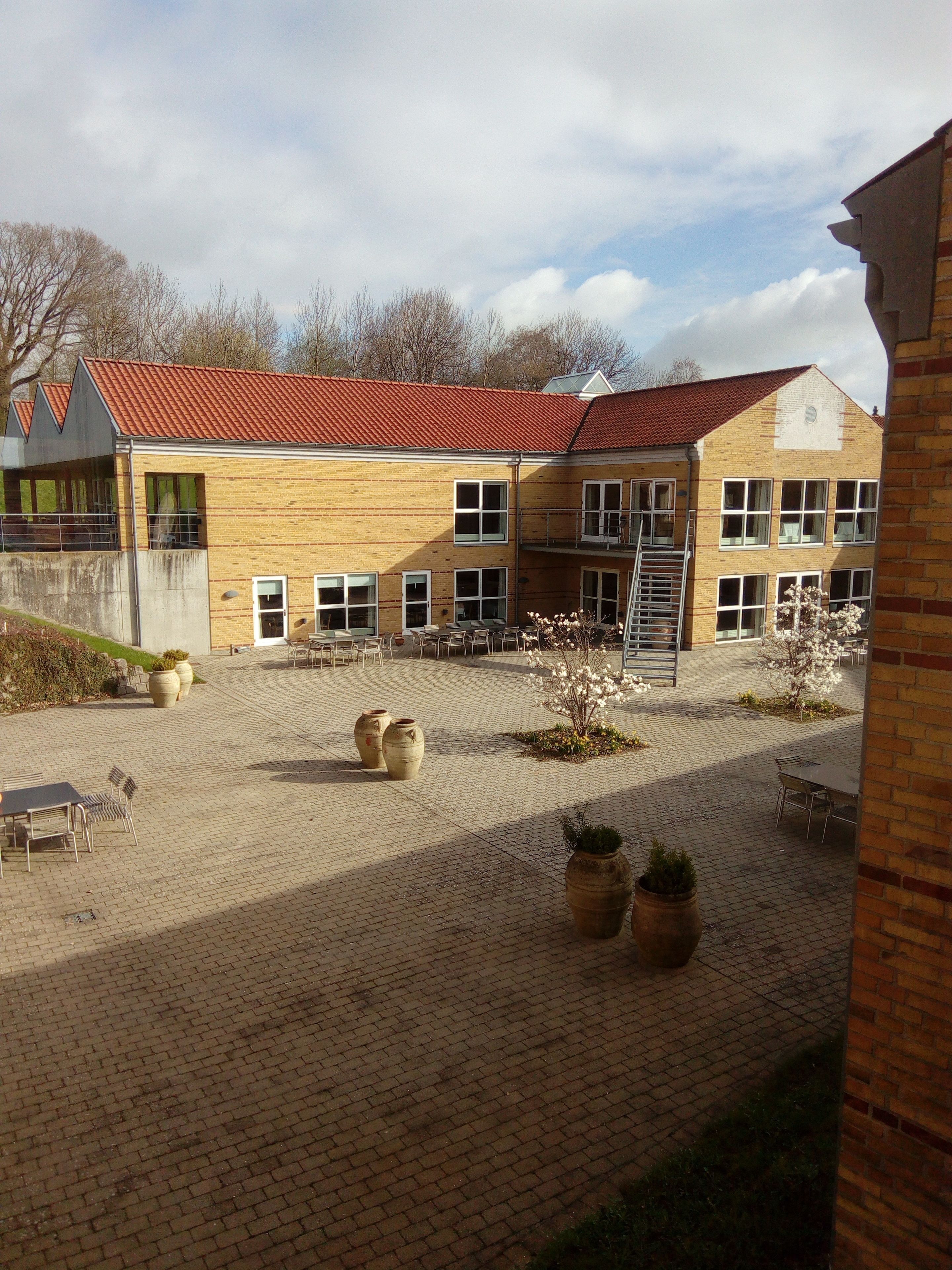 A building with a red-tiled roof, multiple windows, and a paved courtyard with outdoor seating and planters.
