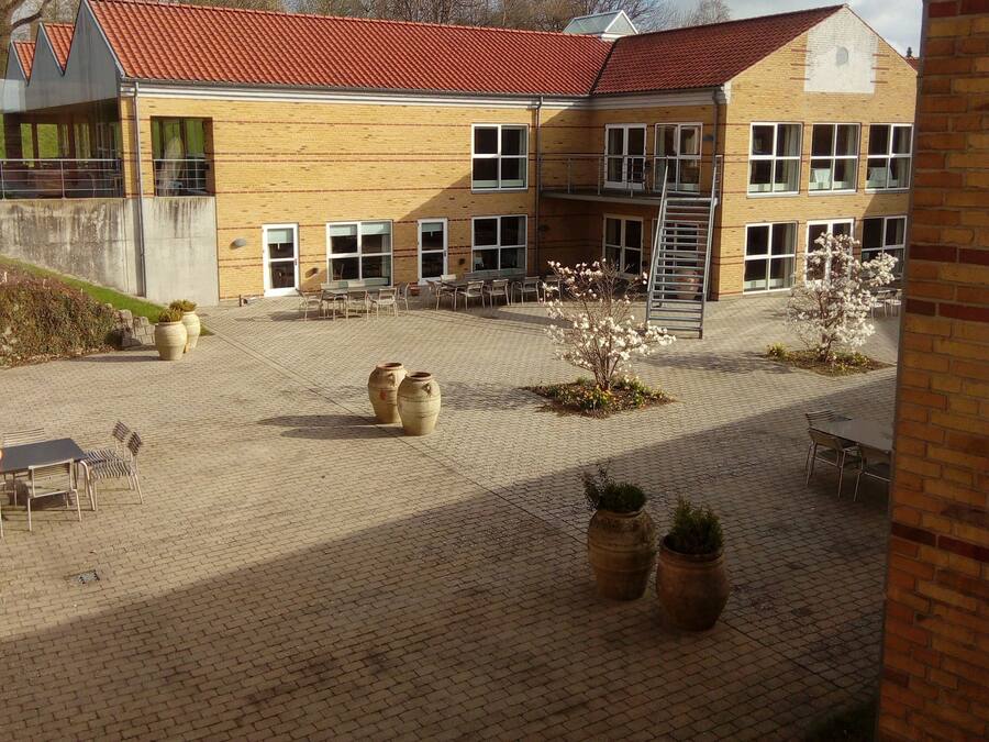 A building with a red-tiled roof, multiple windows, and a paved courtyard with outdoor seating and planters.