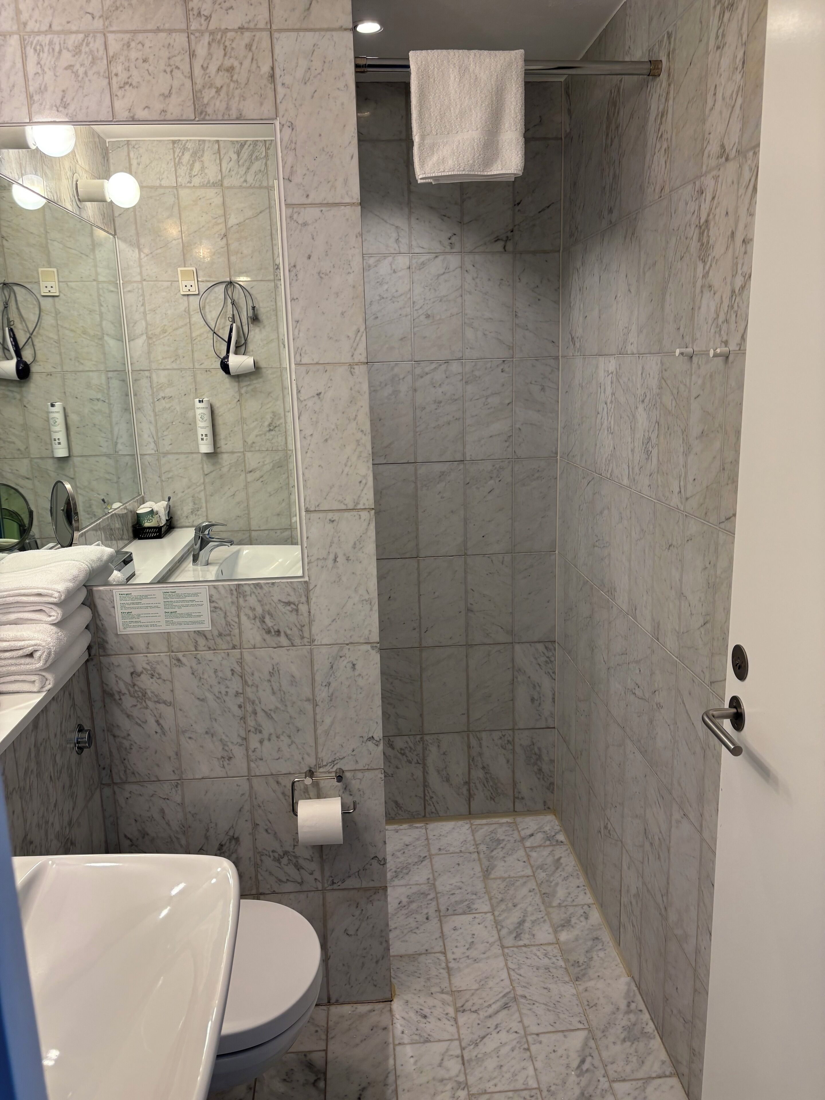 Une salle de bain avec un sol en marbre, un lavabo, des toilettes, une douche avec une serviette suspendue et un miroir avec des lumières.