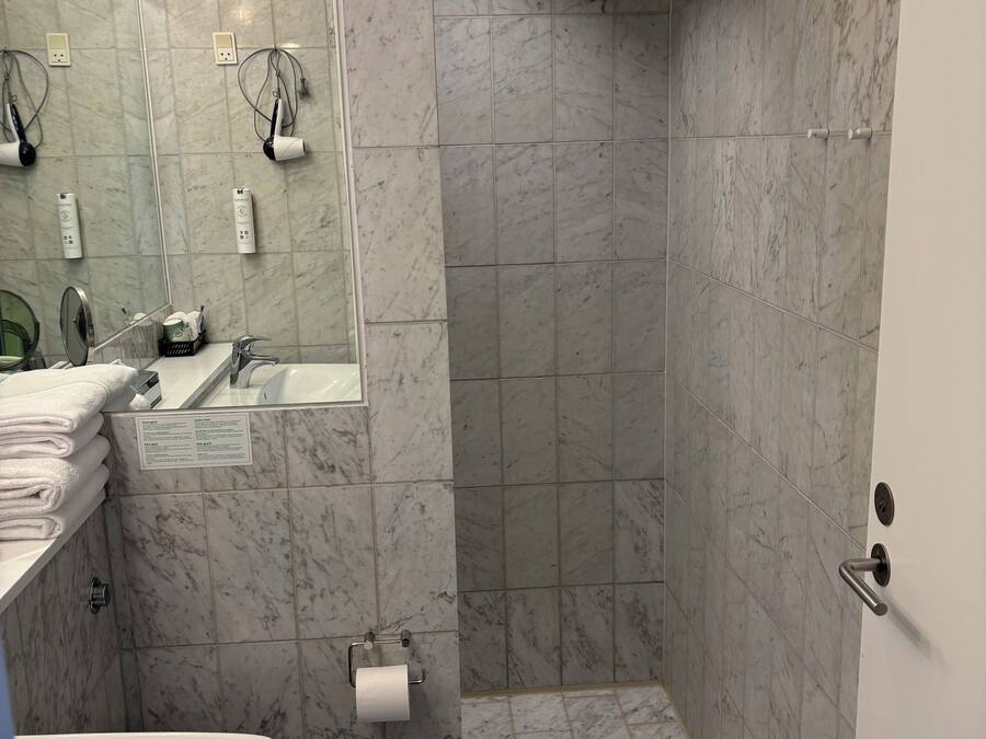 Un baño con piso de mármol, un lavamanos, un inodoro, una ducha con una toalla colgada y un espejo con iluminación.