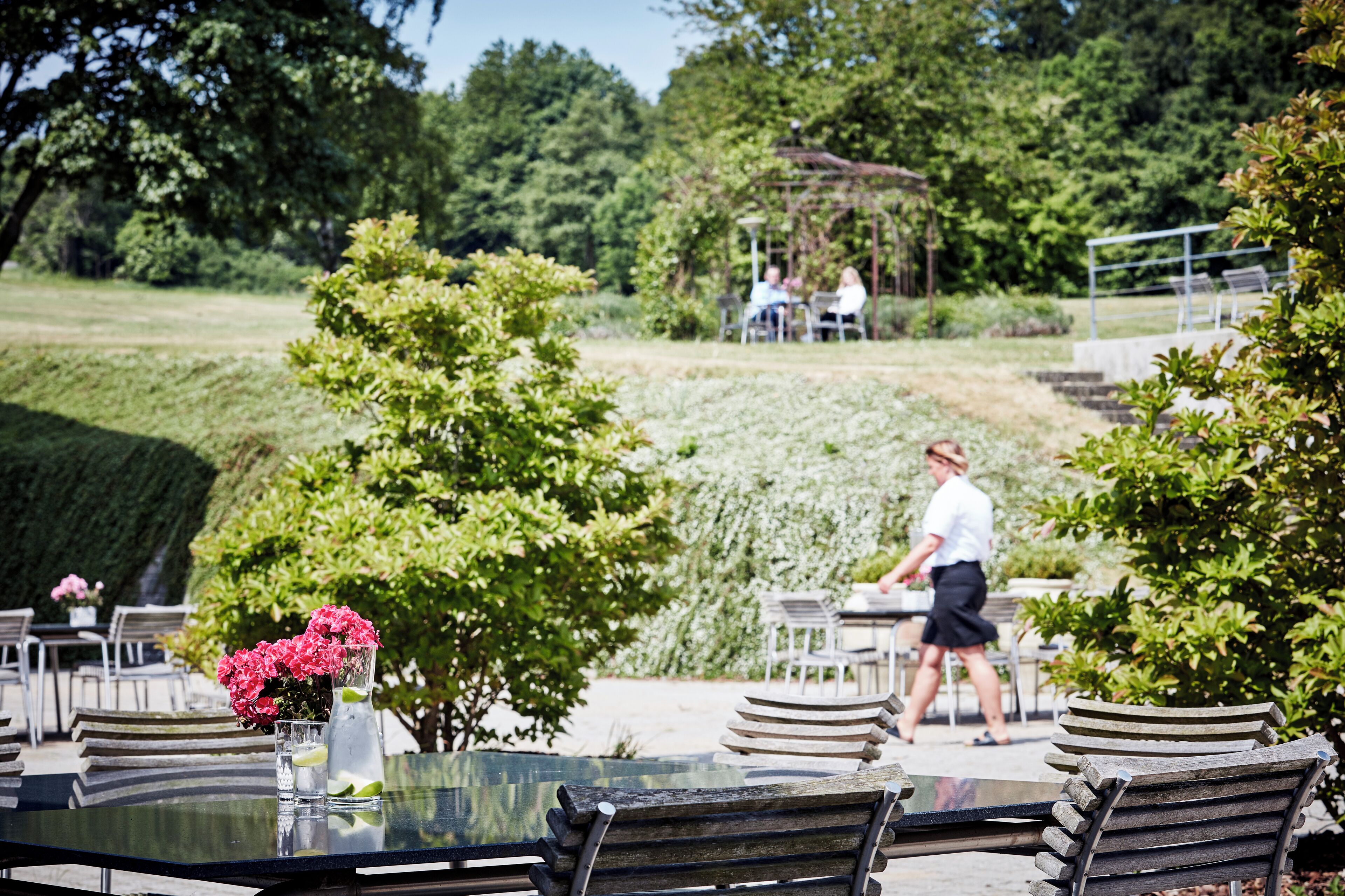 An outdoor dining area with tables and chairs, surrounded by greenery and a walking path.