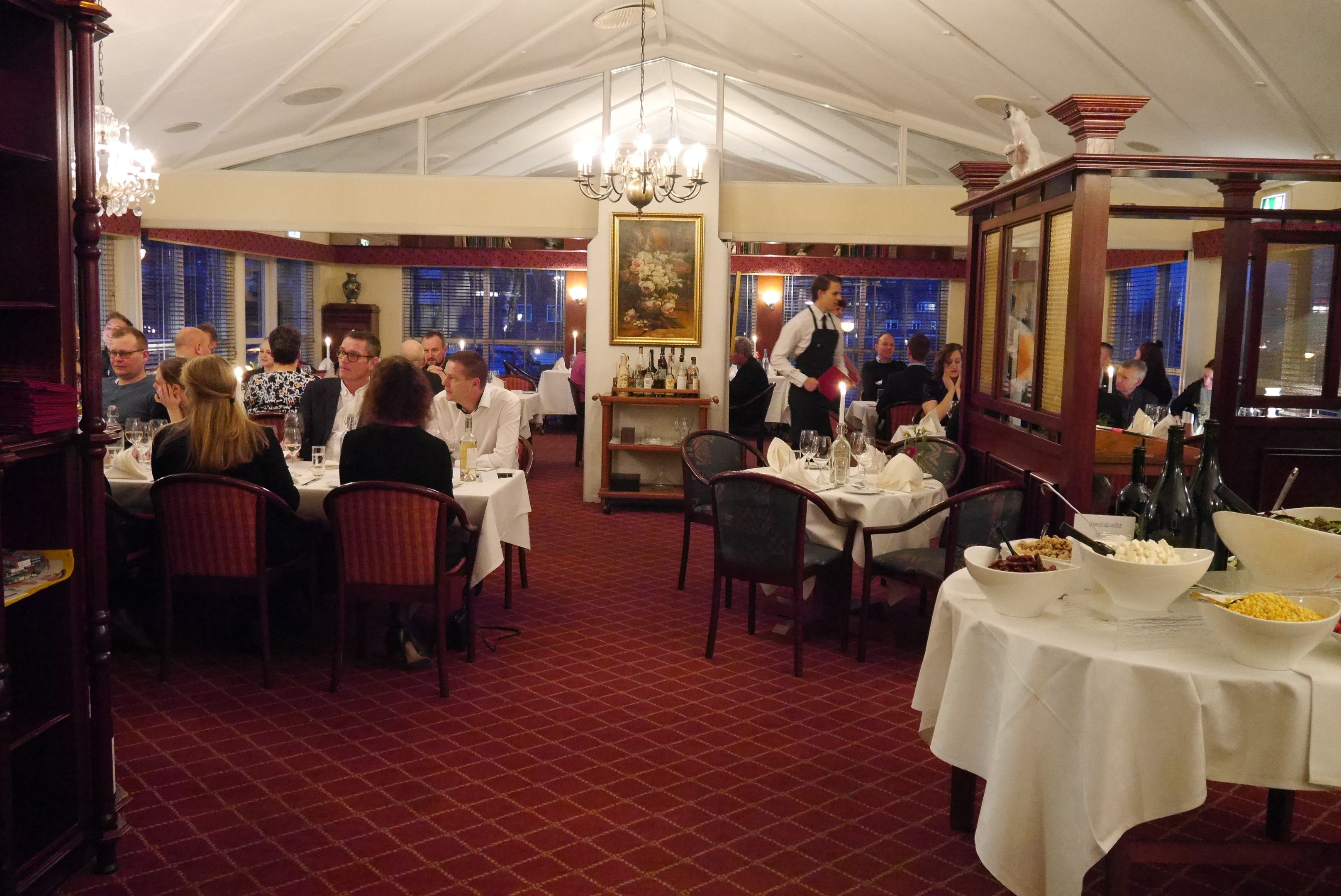A restaurant with tables set for dining, a waiter attending to guests, and a chandelier hanging from the ceiling.