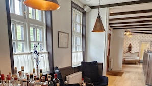 A spacious room with wooden flooring, a black armchair, a wooden table with liquor bottles, and large windows with white curtains.