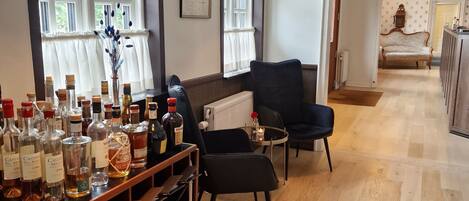 A spacious room with wooden flooring, a black armchair, a wooden table with liquor bottles, and large windows with white curtains.