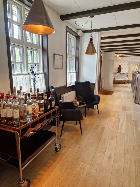 Amplia habitación con suelo de madera, un sillón negro, una mesa de madera con botellas de licor y ventanas grandes con cortinas blancas.