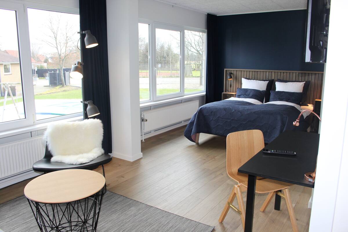 Une chambre moderne avec un lit, un bureau et une chaise. Il y a une fenêtre avec des rideaux et une vue sur l’extérieur.