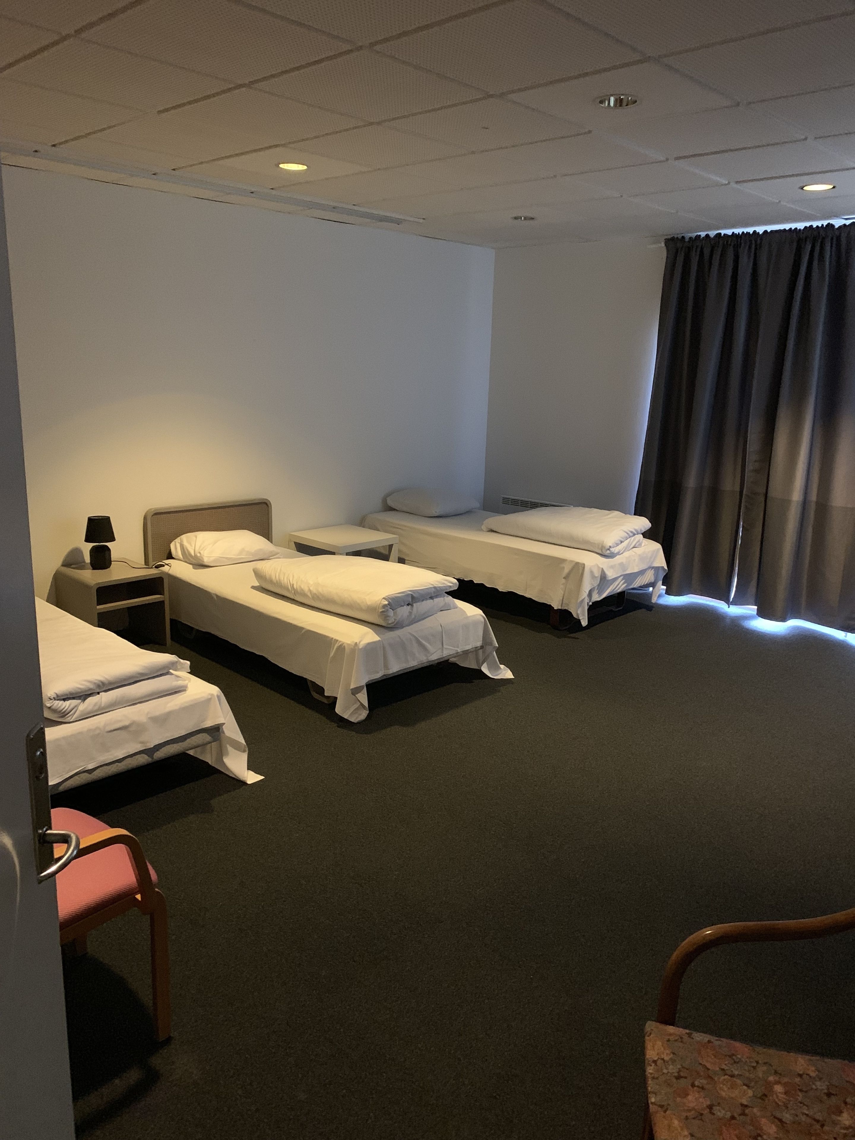 A hotel room with two single beds, a small table with a lamp, a chair, and a window with curtains.