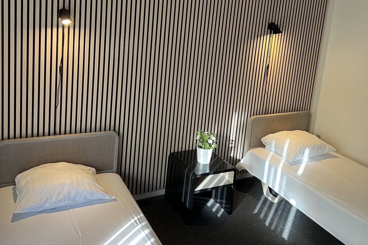 Quarto de hotel com duas camas de solteiro, uma mesinha com uma planta e uma parede com listras verticais.