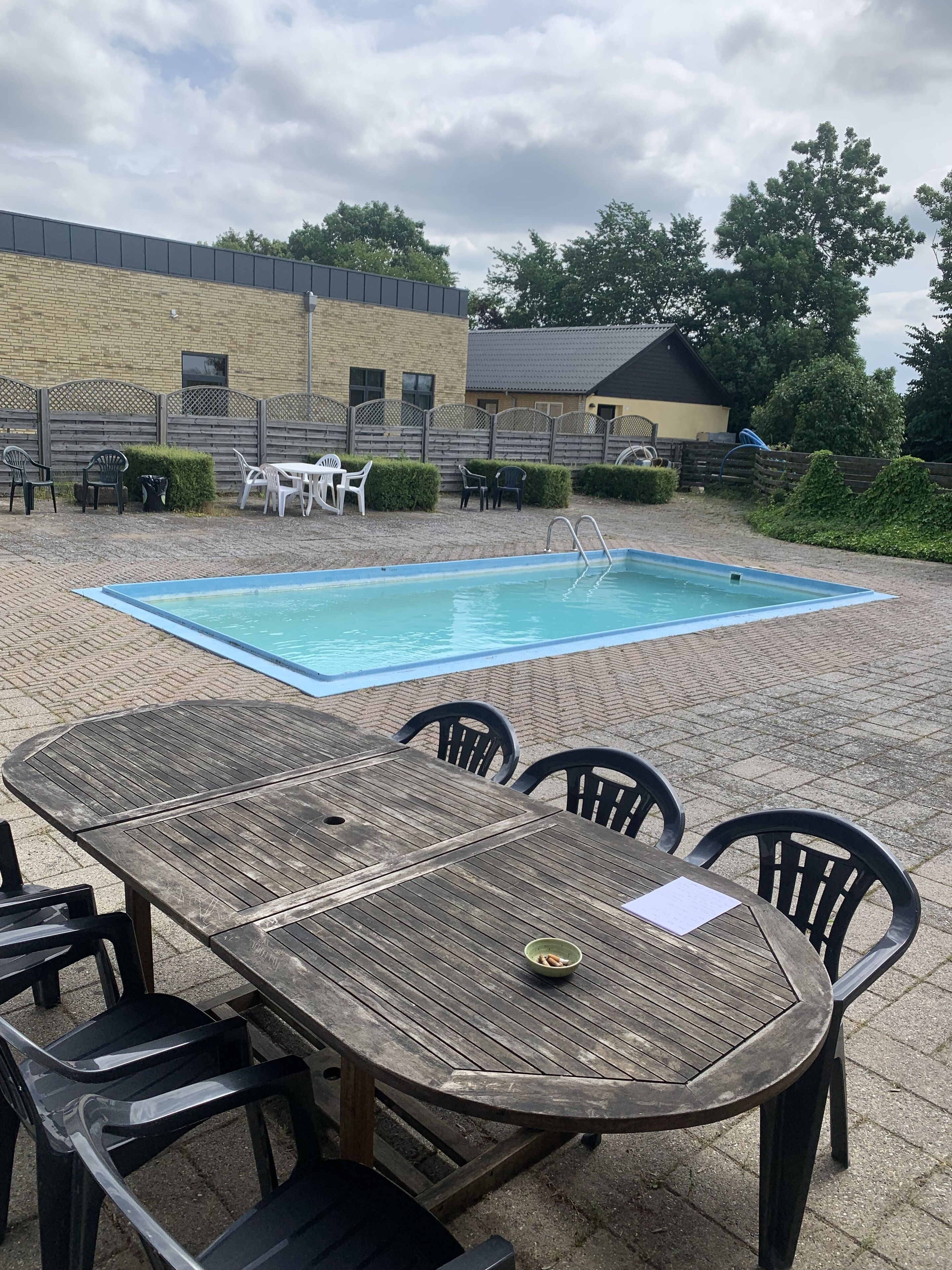 A pool area with a wooden table and chairs, surrounded by a fence and a building in the background.