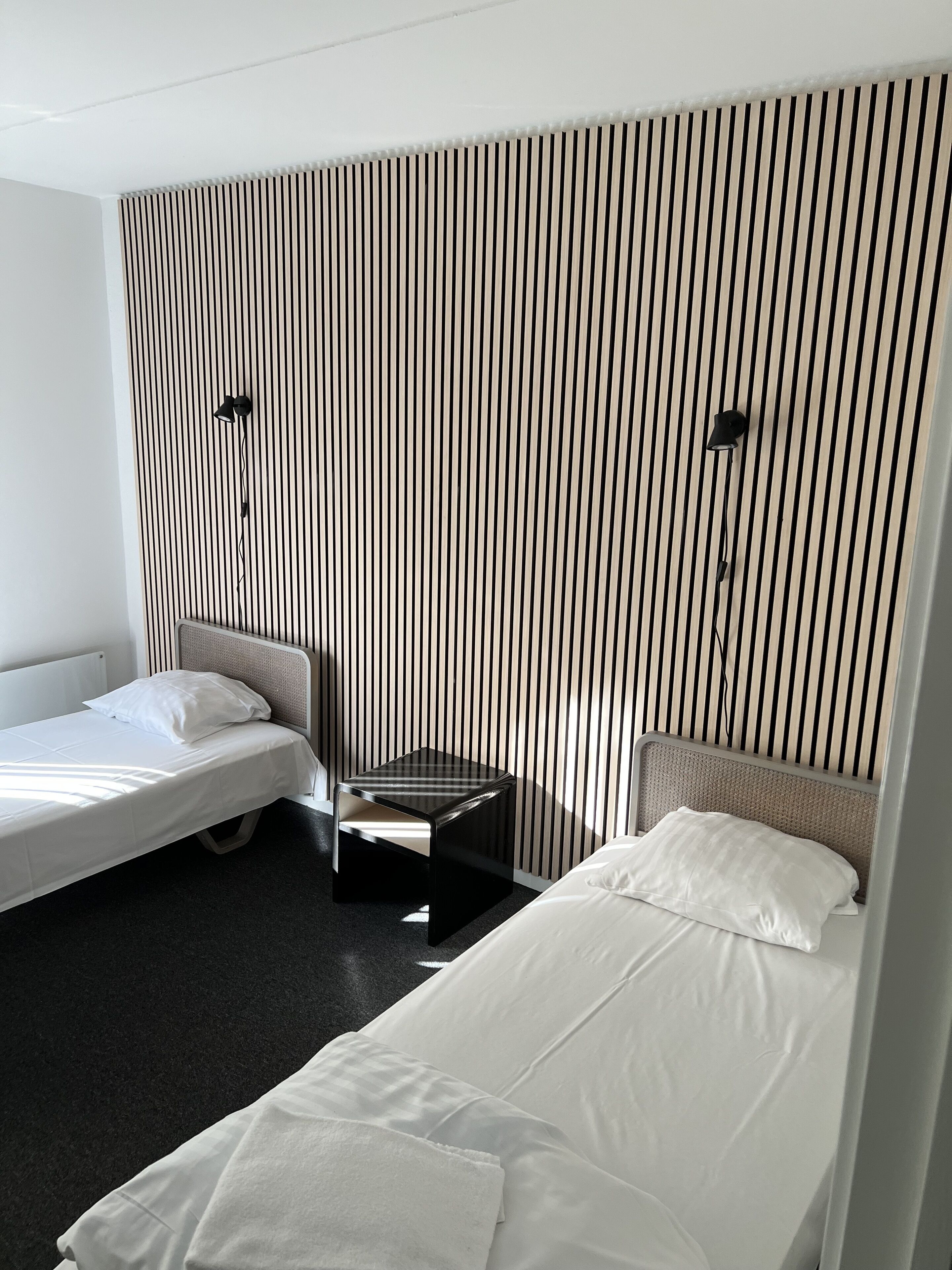Een hotelkamer met twee eenpersoonsbedden, een klein nachtkastje en een wand met verticale strepen.