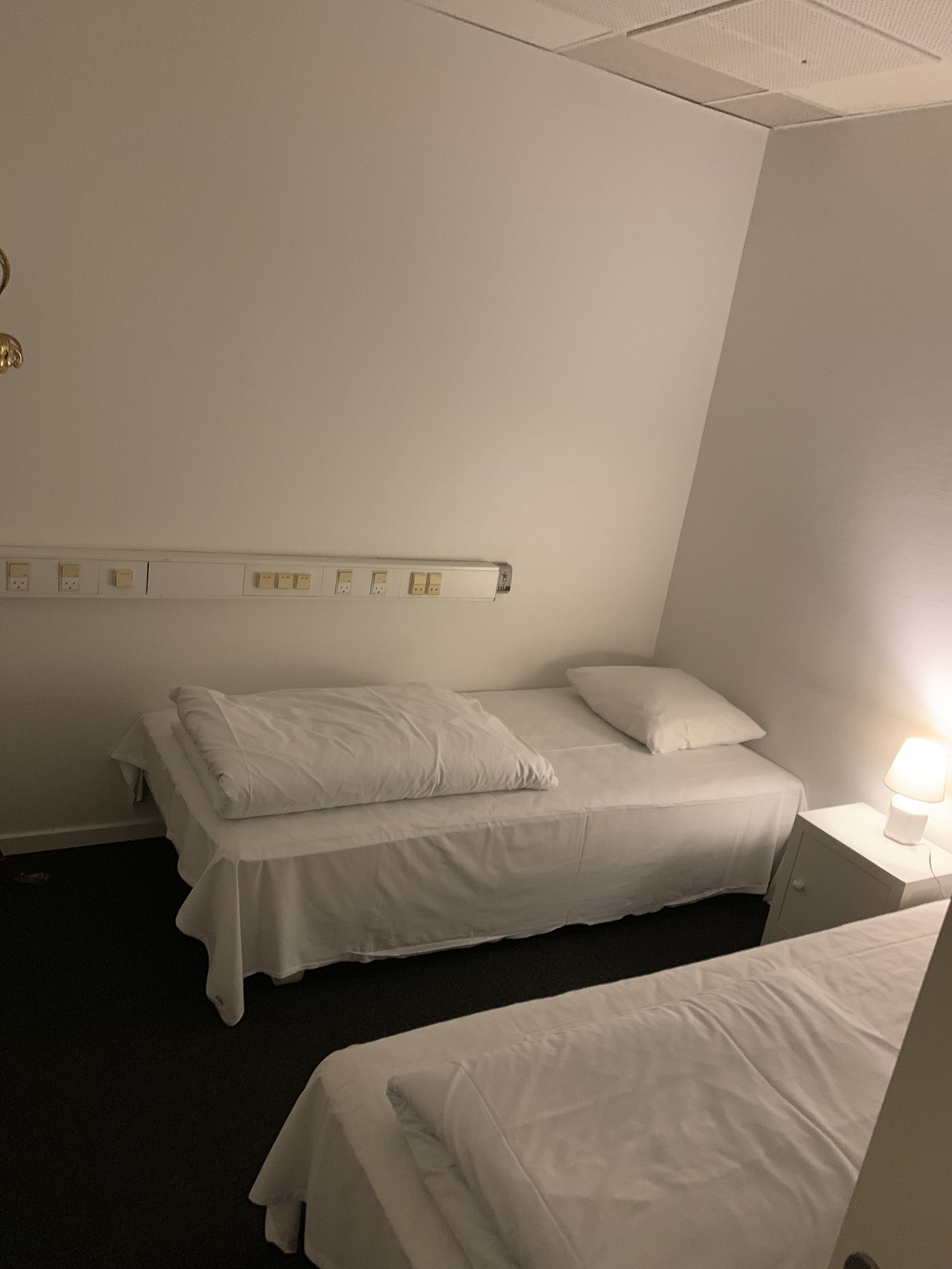 A hotel room with a single bed, a bedside table with a lamp, and a wall-mounted power outlet.