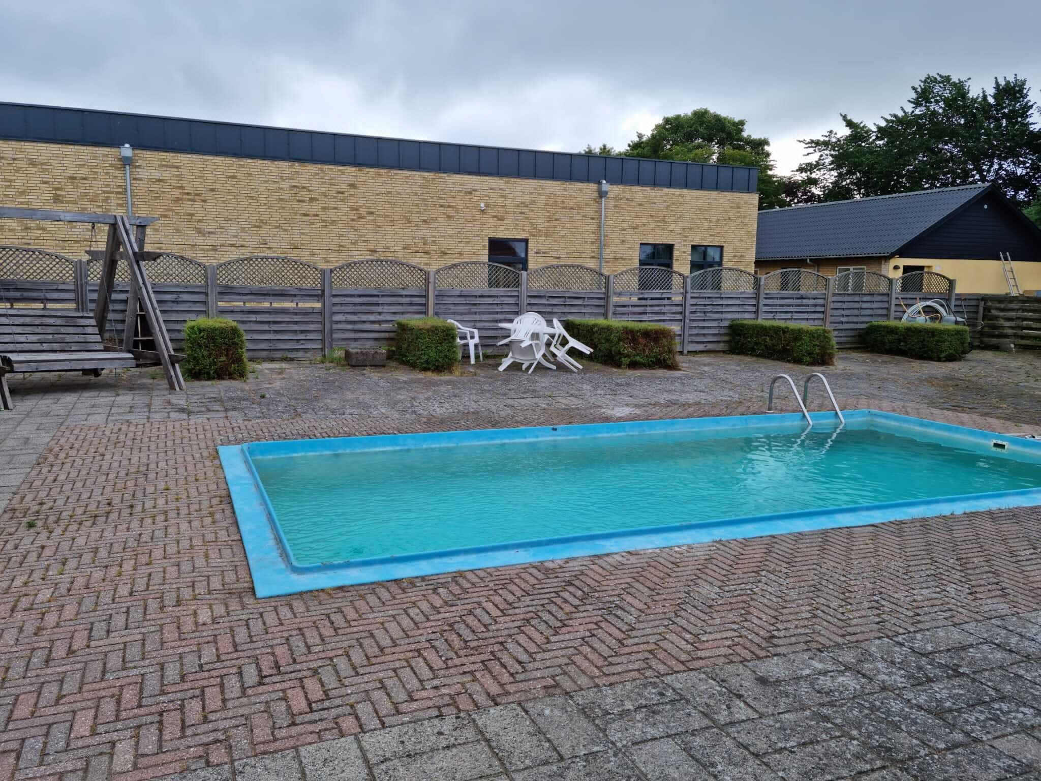 A rectangular swimming pool with a blue edge, surrounded by a brick patio. There are two white chairs and a ladder near the pool.