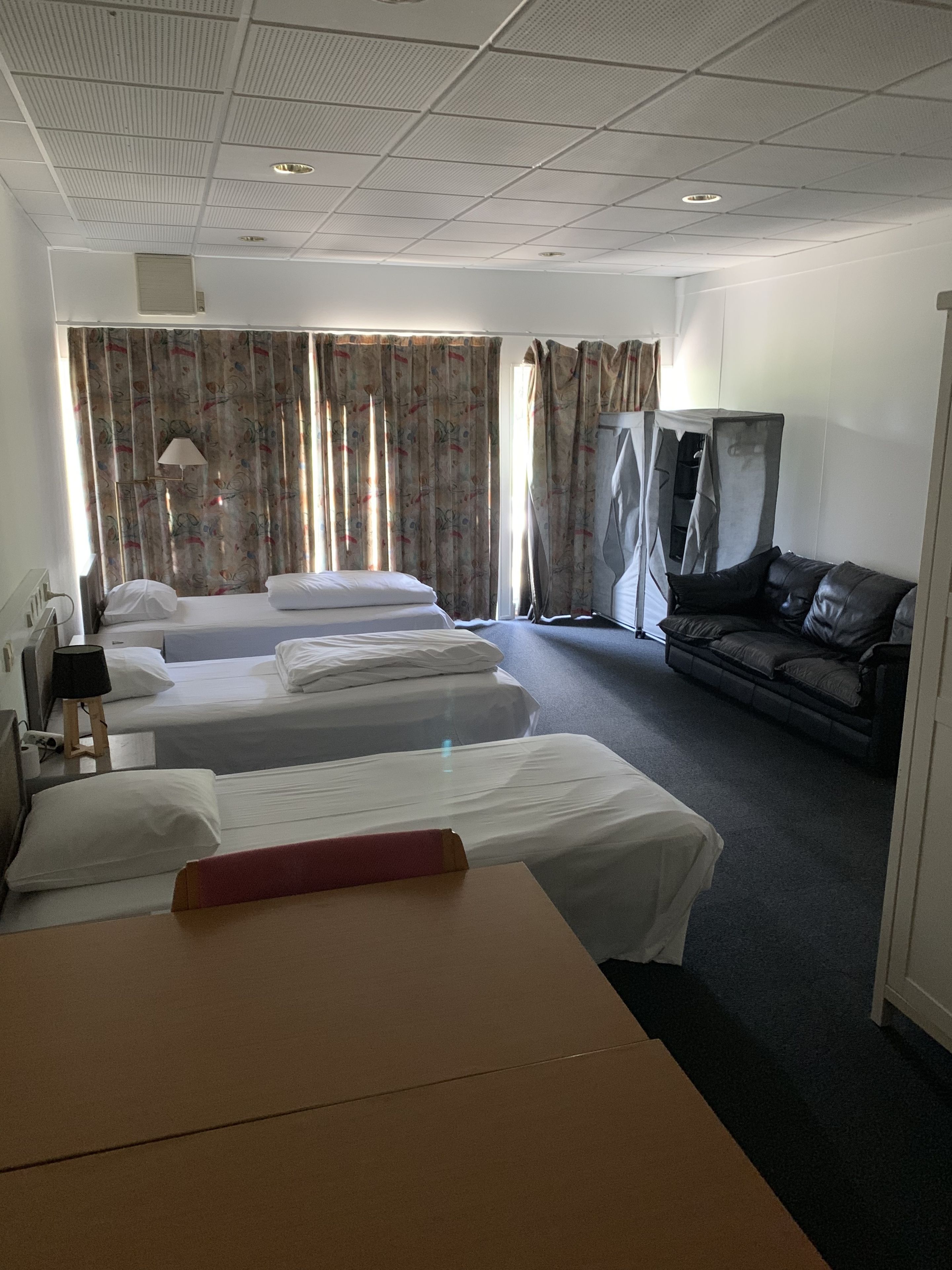 Quarto de hotel com duas camas, um sofá, uma televisão e uma janela grande com cortinas.
