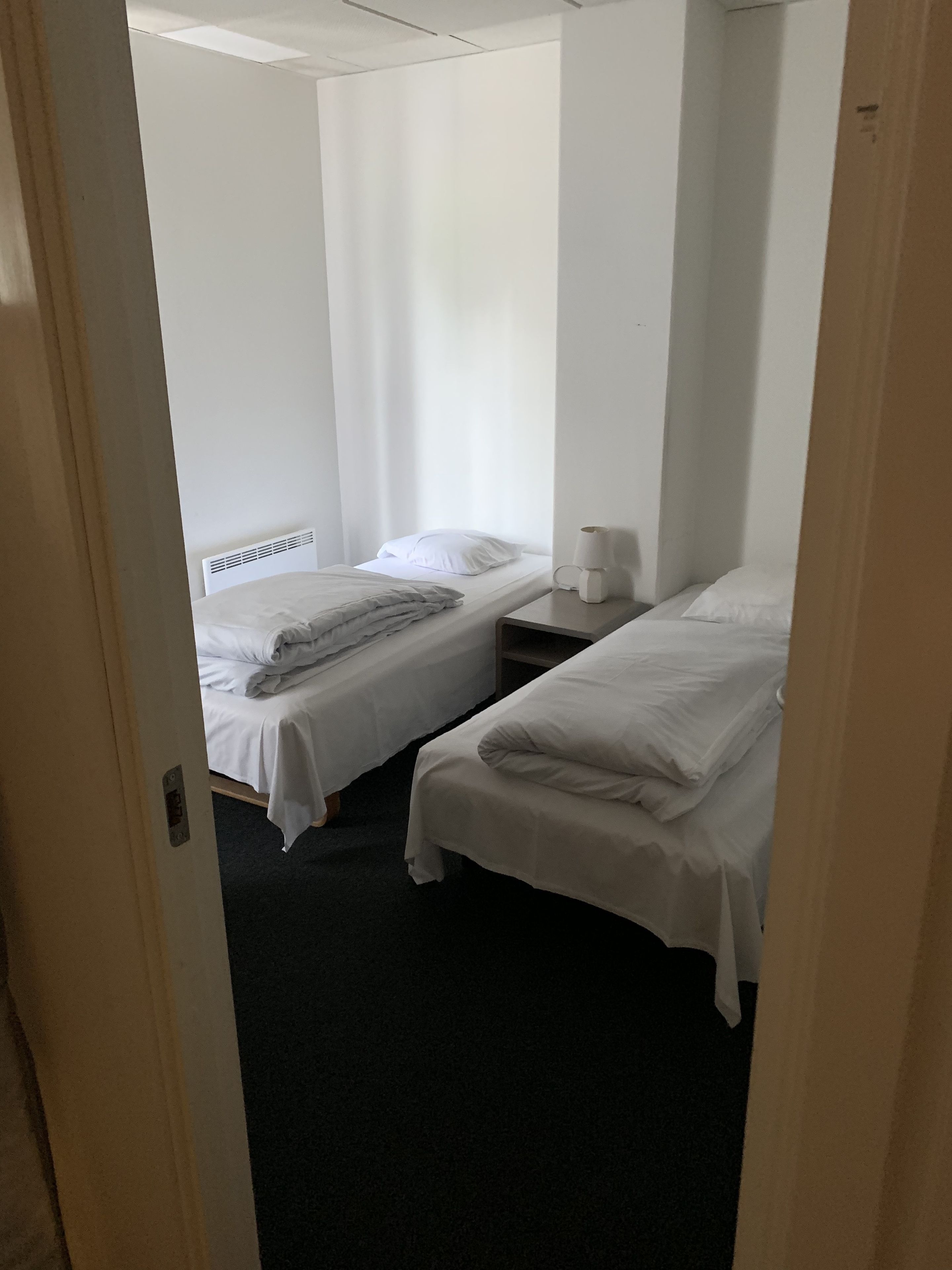 Habitación de hotel con dos camas individuales, una mesita de noche con lámpara y una ventana con cortinas translúcidas.