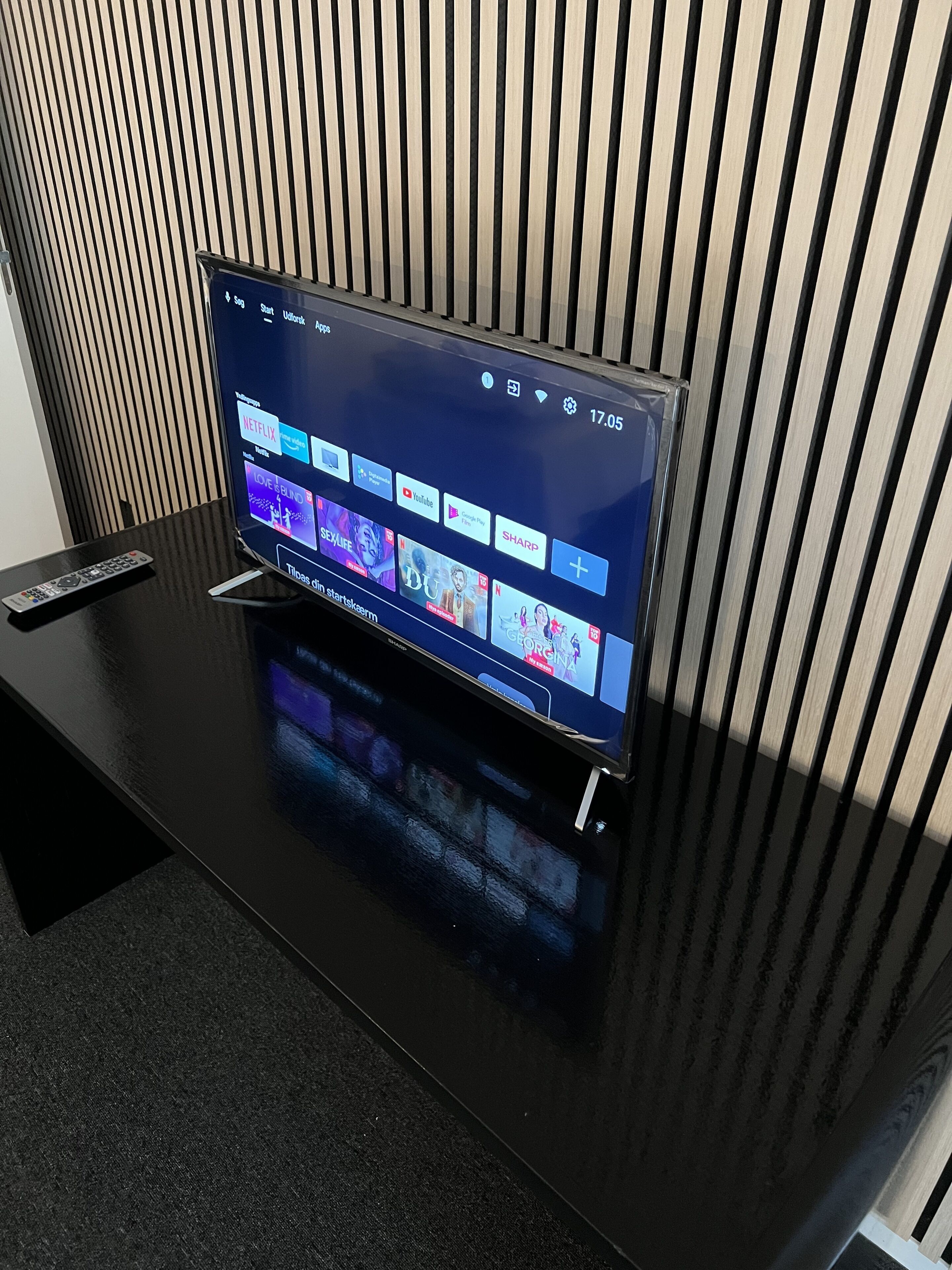 A flat-screen TV on a black stand with a remote control beside it, displaying a streaming service interface.
