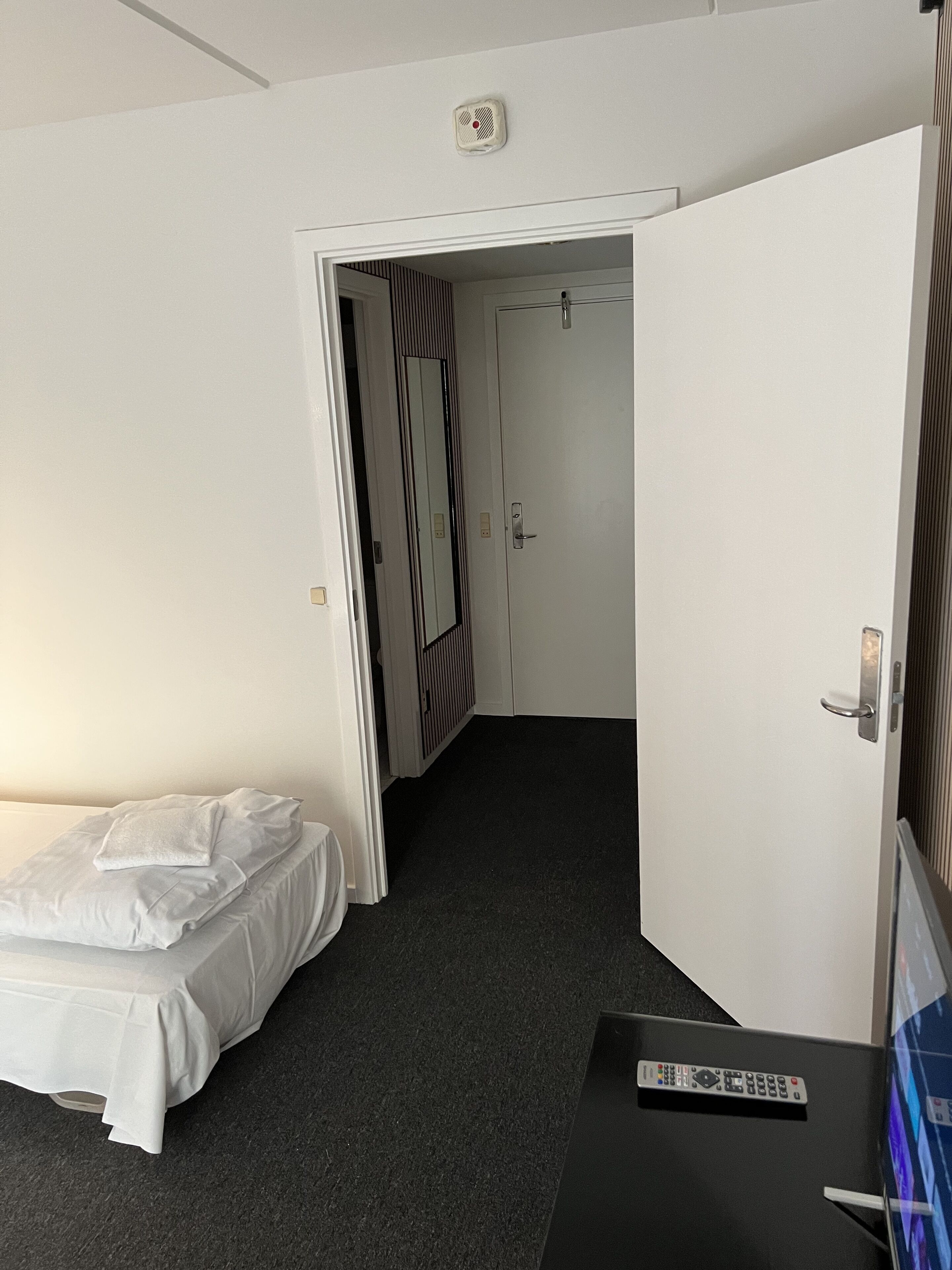 A hotel room with a bed, a desk with a remote control, a door leading to another room, and a smoke detector on the ceiling.