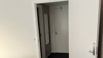 A hotel room with a bed, a desk with a remote control, a door leading to another room, and a smoke detector on the ceiling.