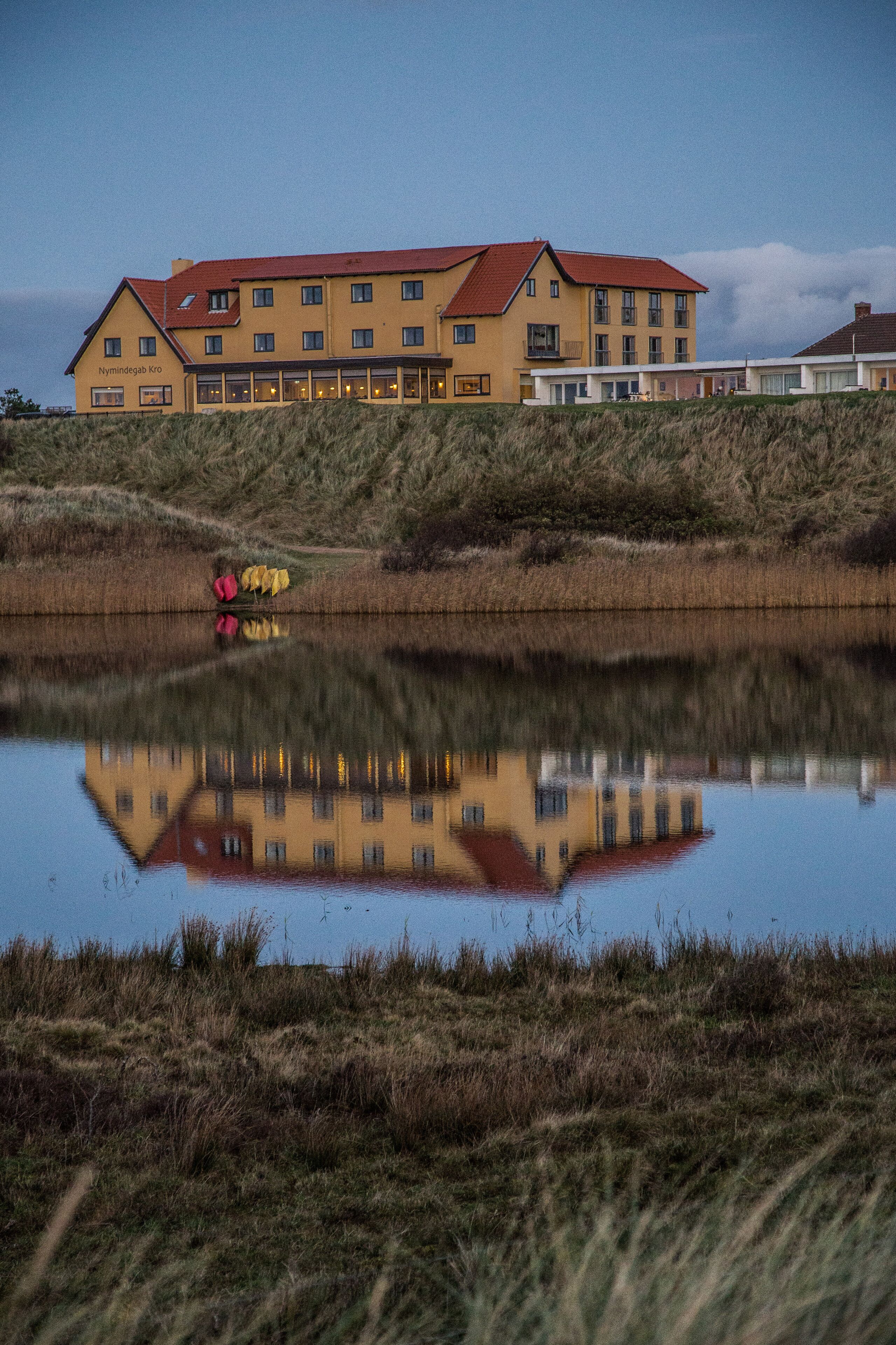 Un edificio grande con techo rojo y varias ventanas, situado junto a una masa de agua que refleja su estructura.