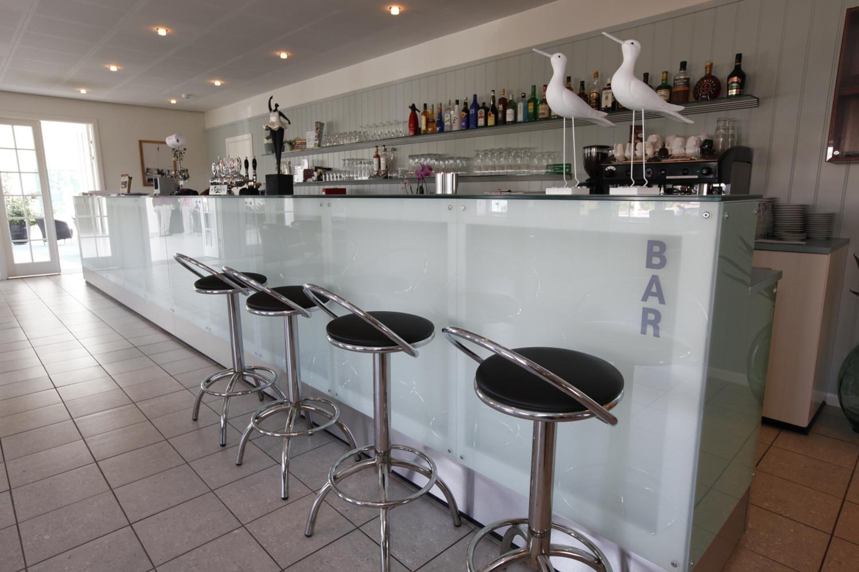 Una zona de bar con mostrador de cristal, taburetes y estantes con botellas y vasos.