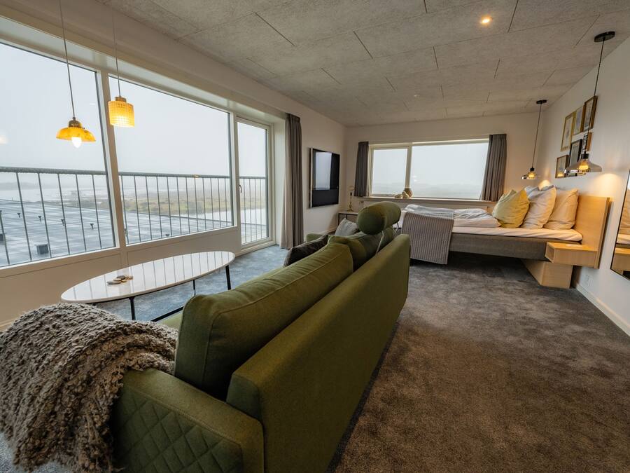 Ein modernes Hotelzimmer mit großem Fenster zum Meer, einem gemütlichen grünen Sofa, einem kleinen Tisch und einem Bett mit Aussicht.