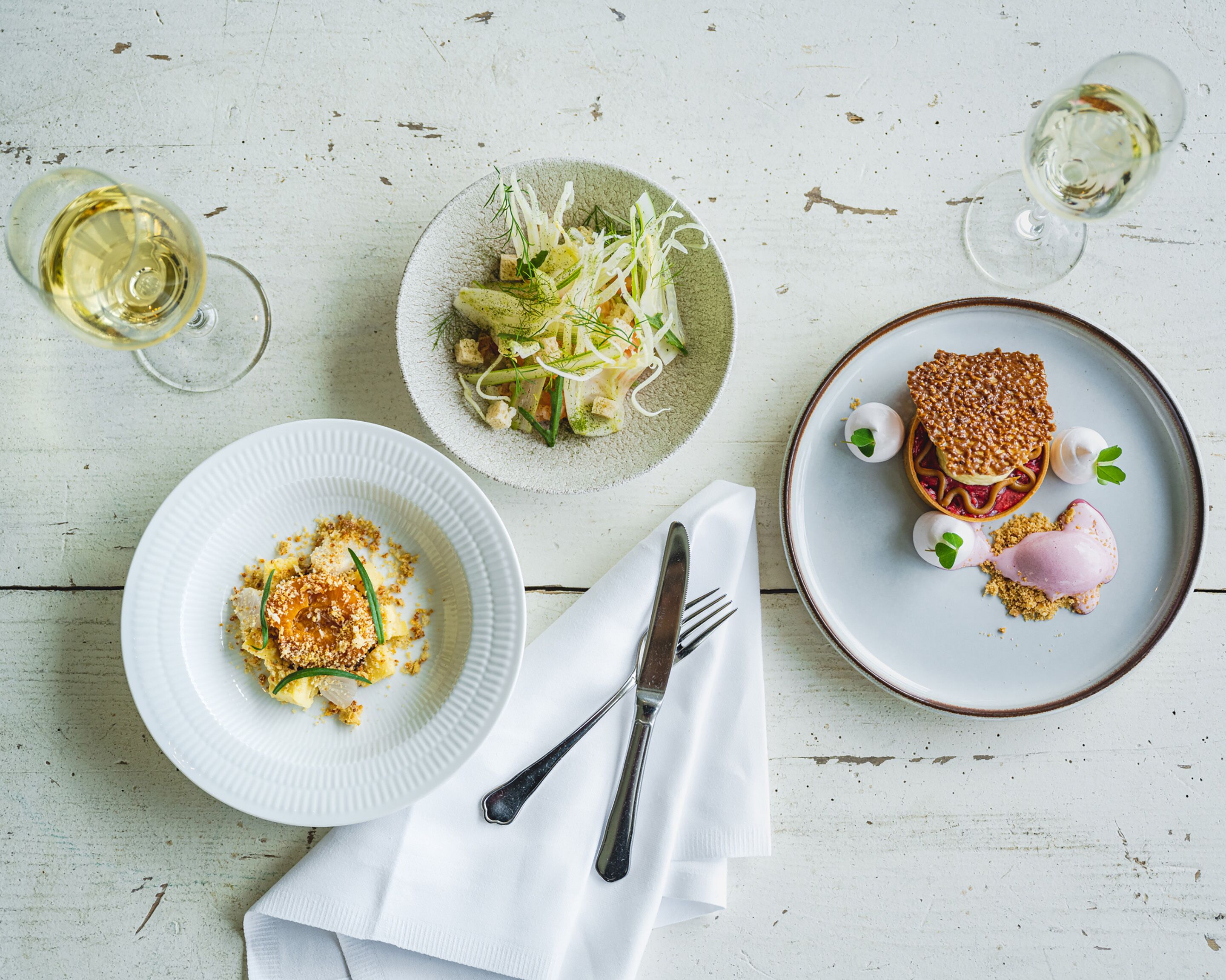 A dining table with three plates of food, two wine glasses, and cutlery on a rustic wooden surface.