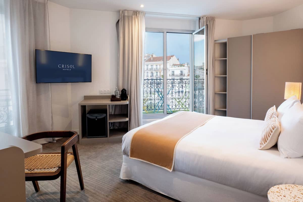 A hotel room with a bed, a desk, a TV, and a view of buildings through a window.