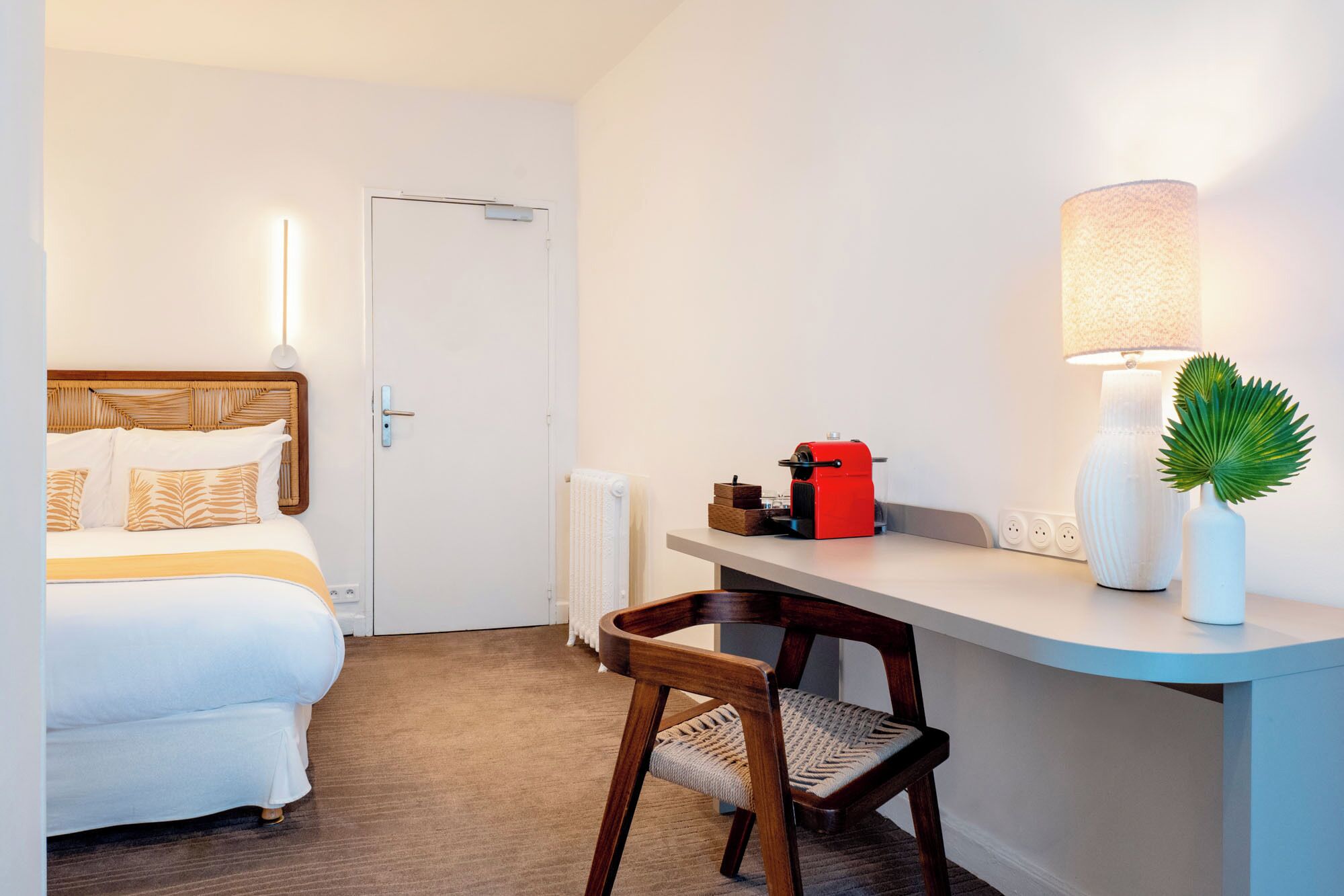 A hotel room with a bed, a desk, a chair, a lamp, a radiator, and a small plant.