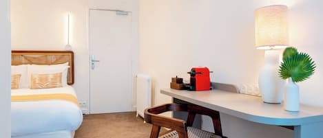 A hotel room with a bed, a desk, a chair, a lamp, a radiator, and a small plant.